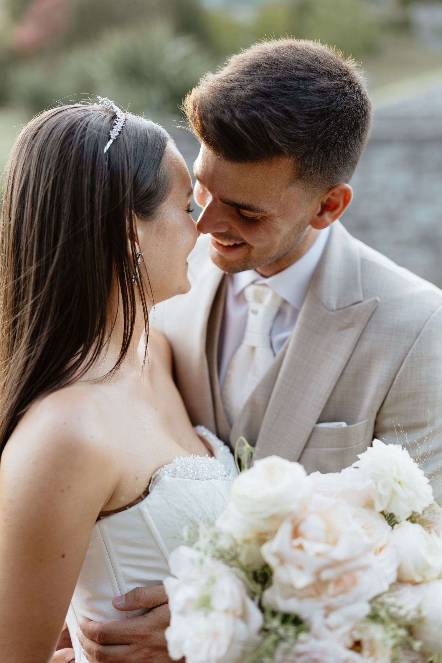 Bride in white dress holds pastel bouquet while sharing tender moment with groom in beige suit.