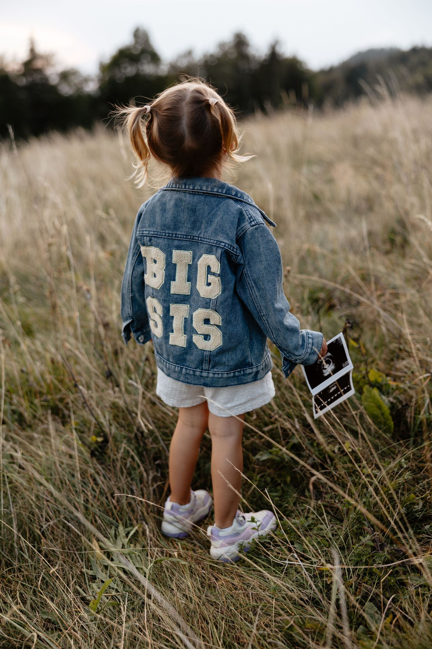 A small child wearing a 'Big Sis' denim jacket holds an ultrasound photo in a grassy field.