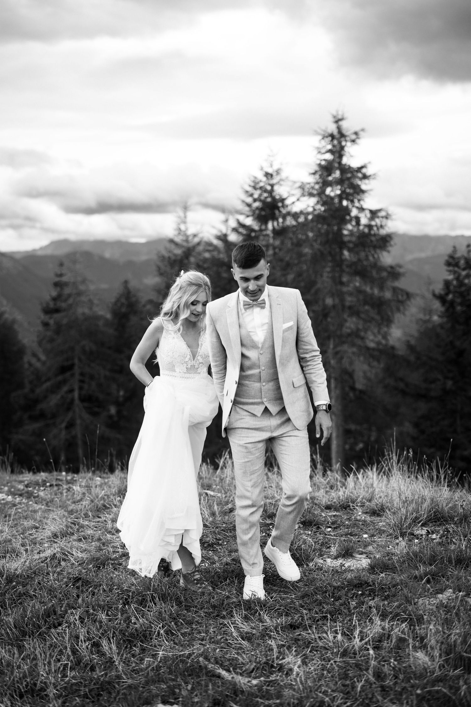 A couple in wedding attire walks together on a scenic mountain hillside with evergreen trees in the background.