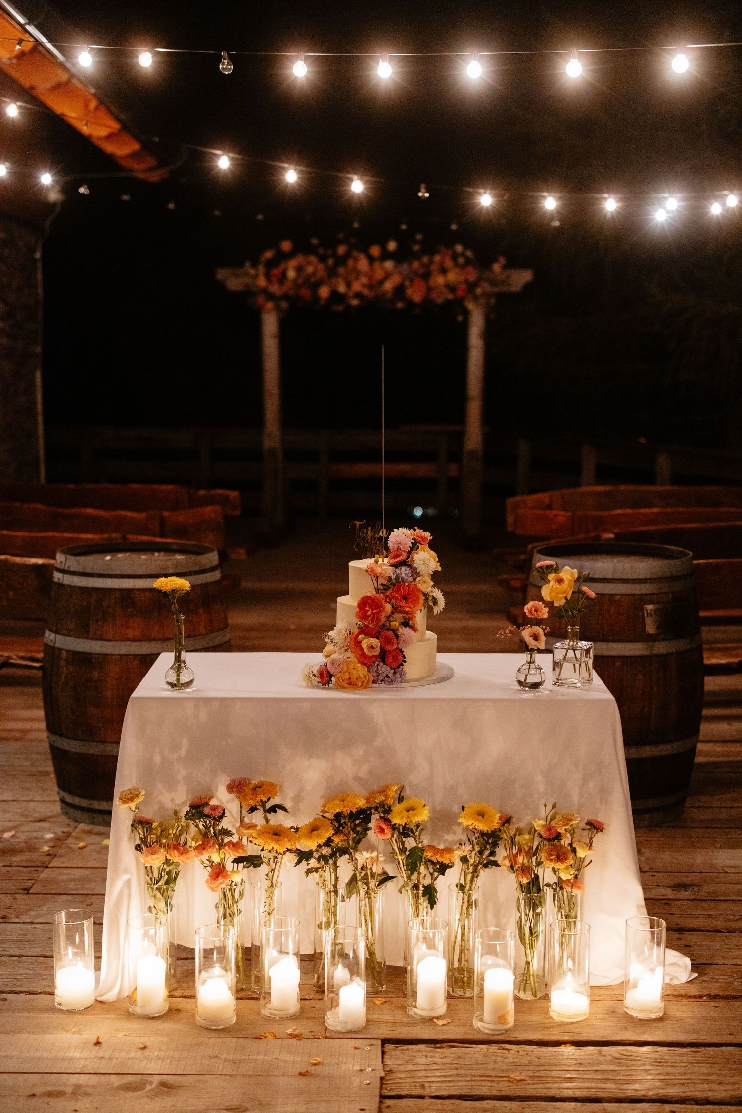 Rustic outdoor wedding reception table decorated with glowing candles and autumn floral arrangements against wooden barrels.