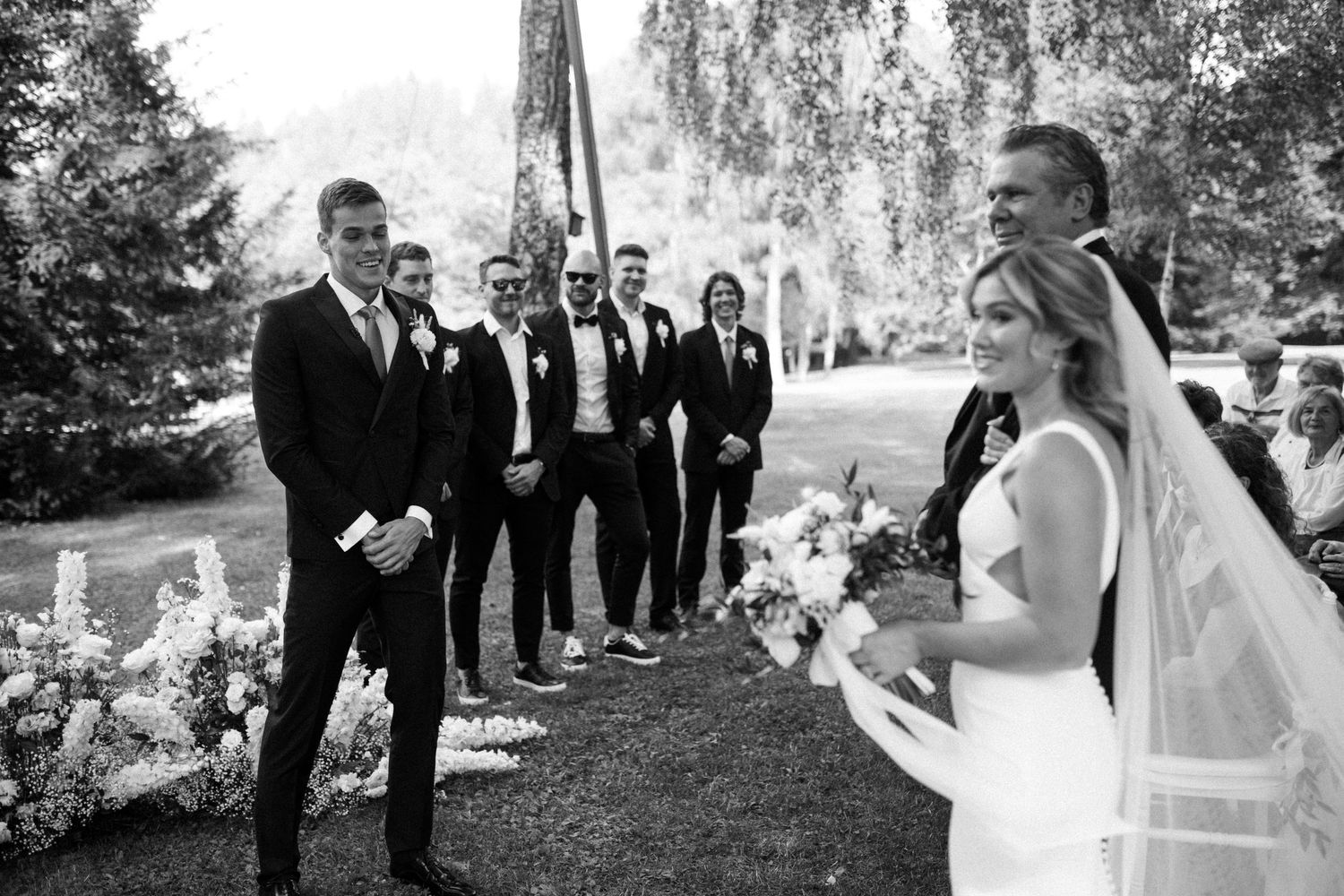 Black and white wedding photo showing bridal party walking through a tree-lined path outdoors.