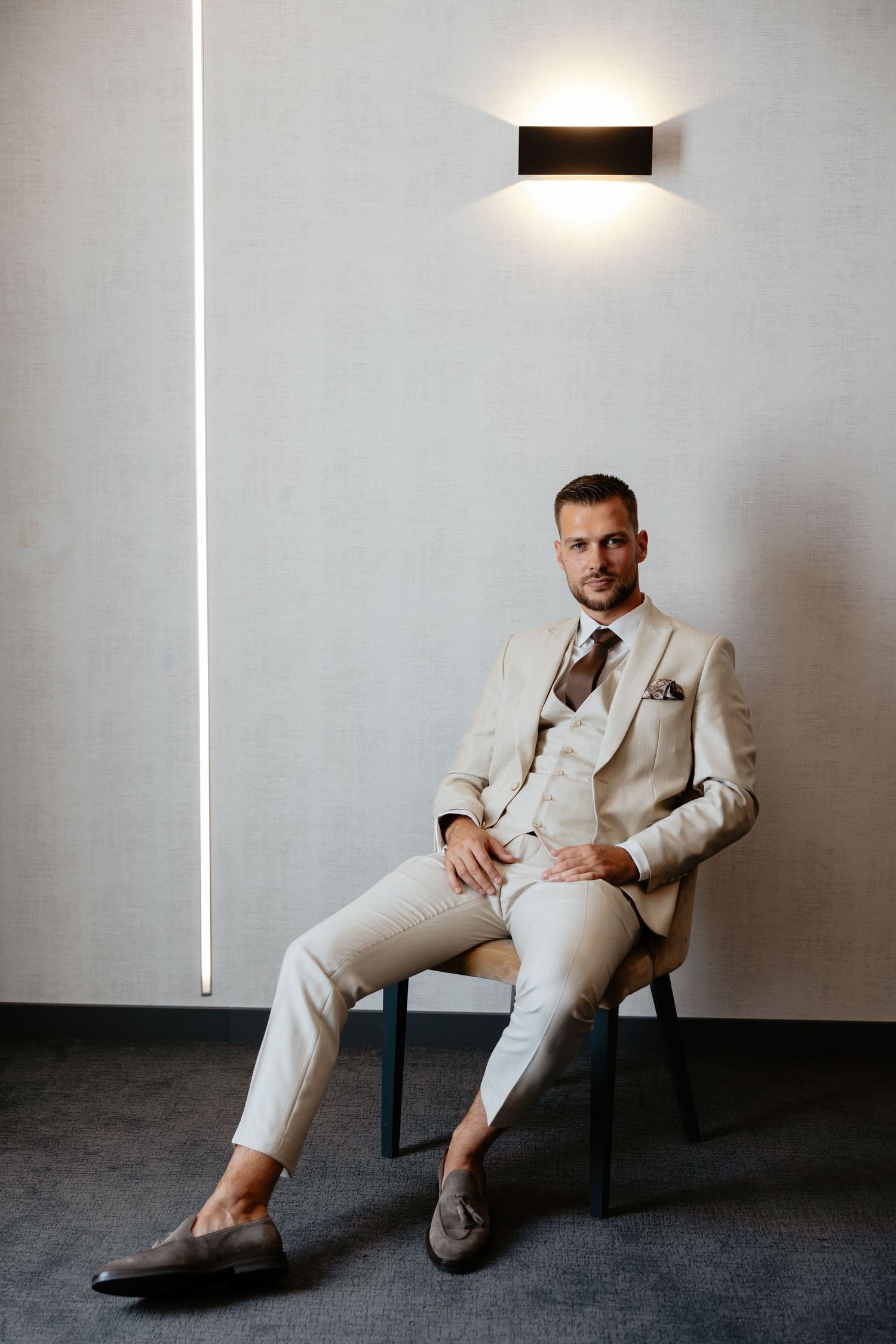 Stylish groom poses in cream colored suit against minimalist grey wall and dark floor.