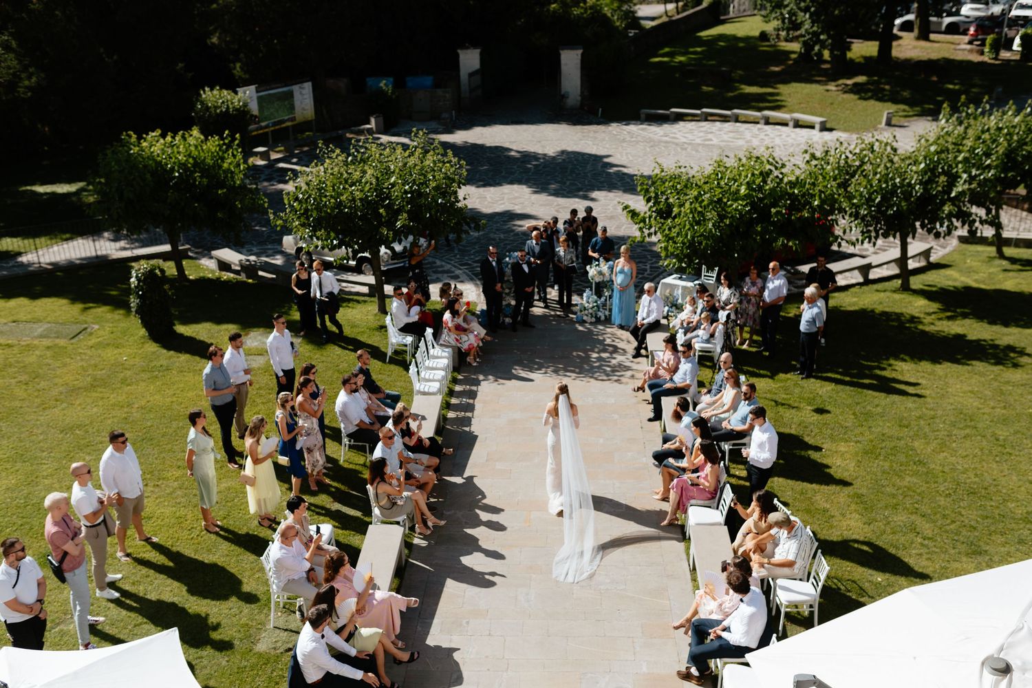 Wedding guests gather outdoors on a sunny day in a garden setting.