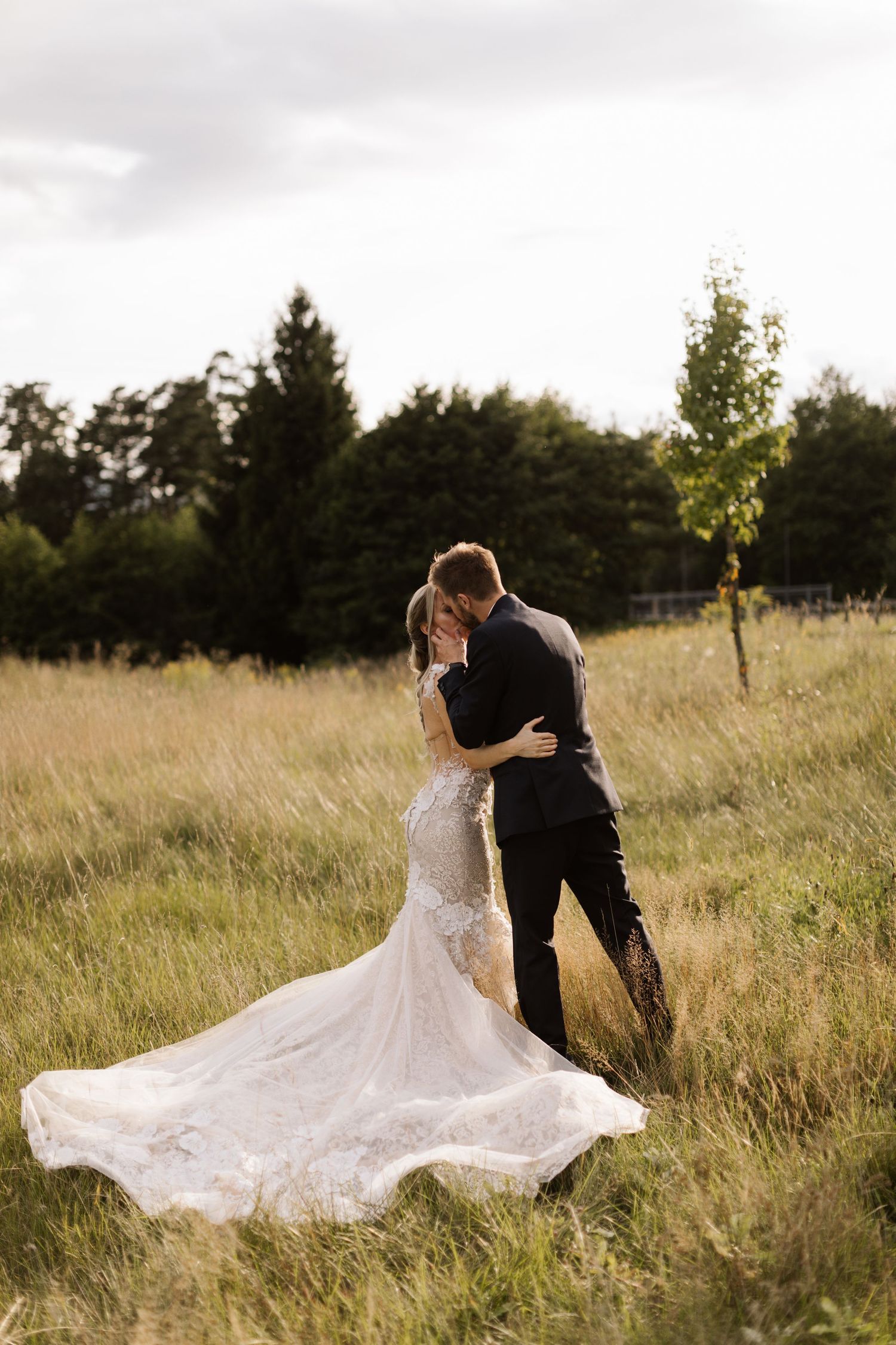 A bride's lace dress flows dramatically as the couple shares an intimate moment in a meadow.