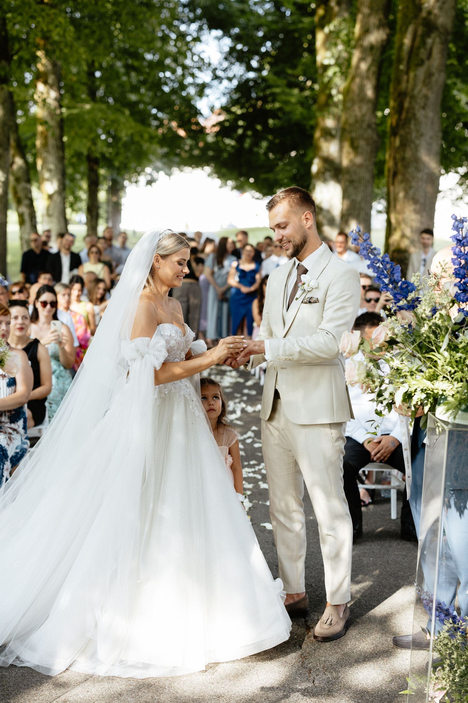 A wedding ceremony in a tree-lined outdoor venue with blue floral decorations.