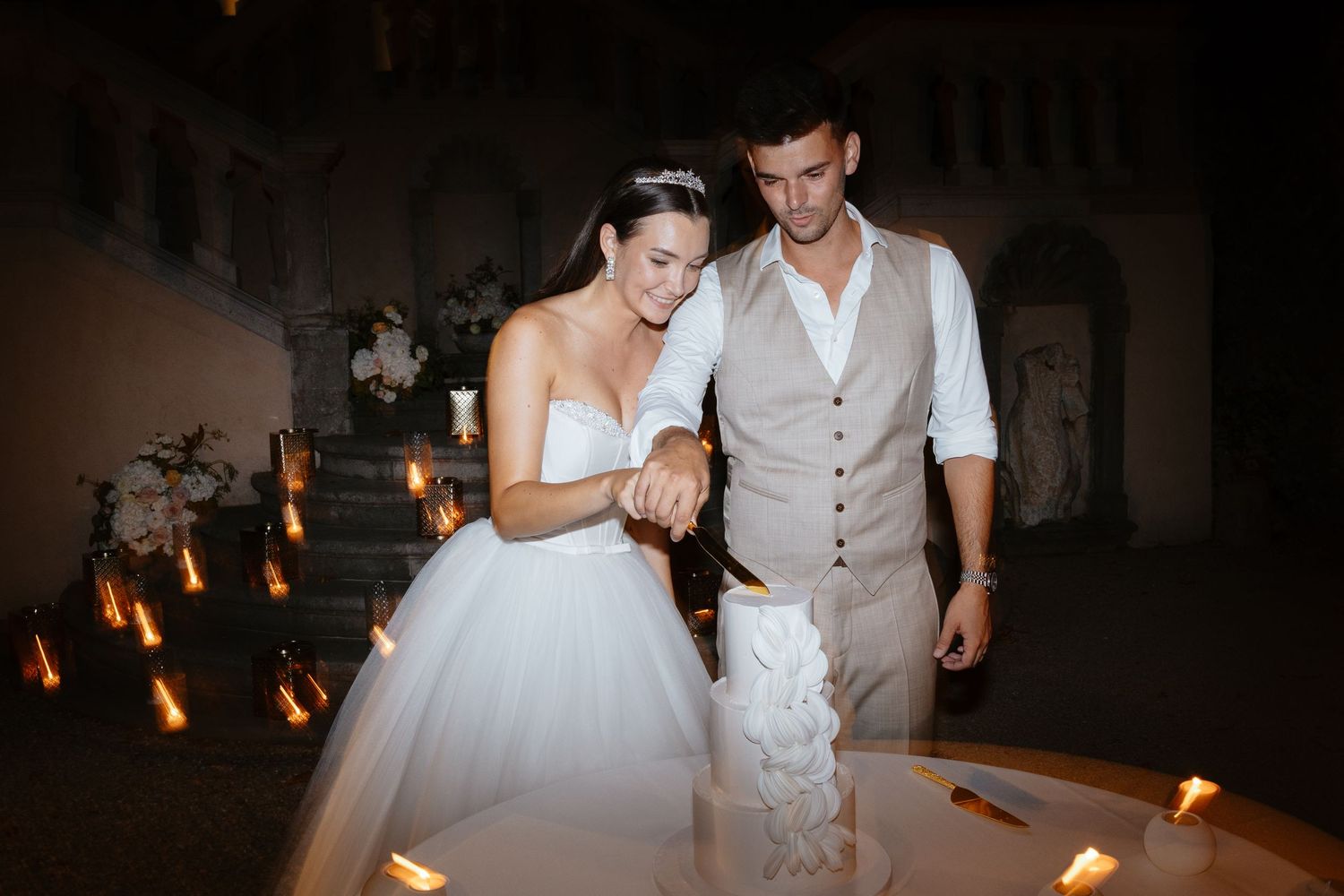 A couple cuts their white wedding cake together in evening lighting.