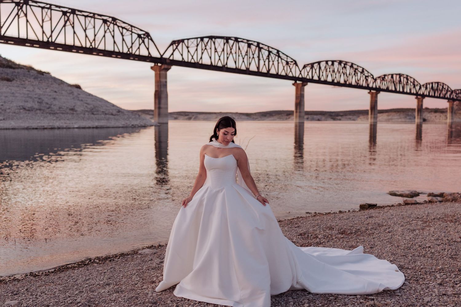 A scenic wedding photoshoot by a river at sunset with a bridge and mountains in the background.