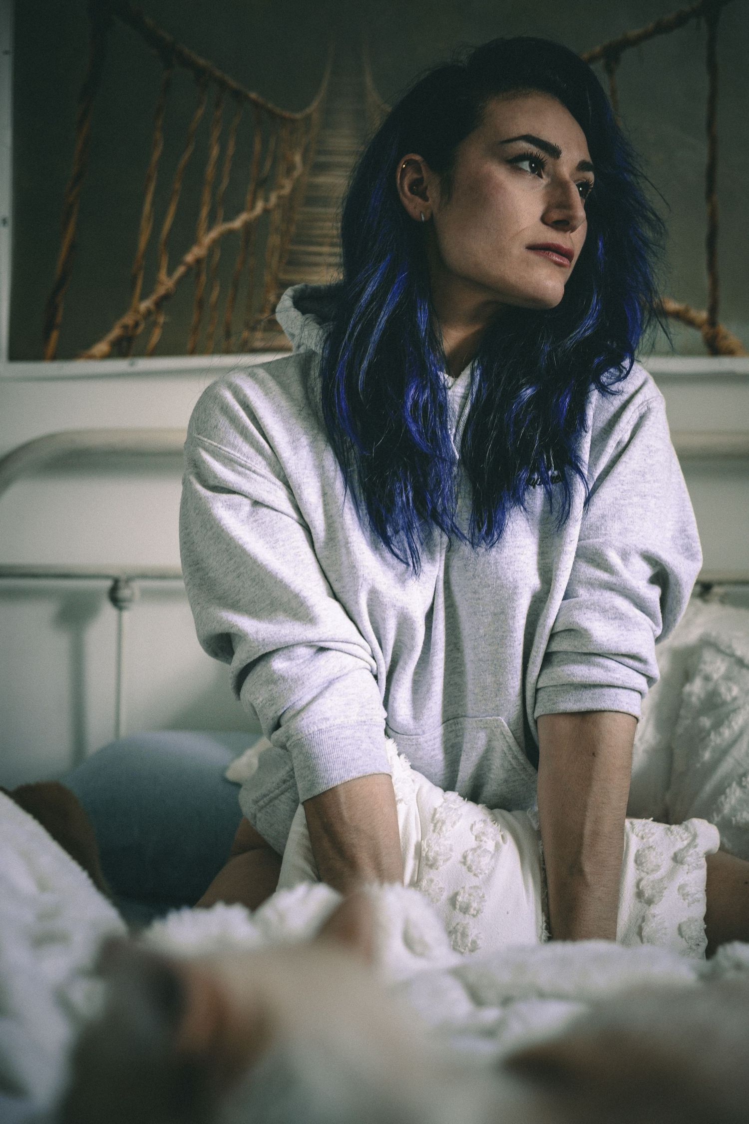 Person in grey hoodie sitting on white bedding with vibrant blue hair.