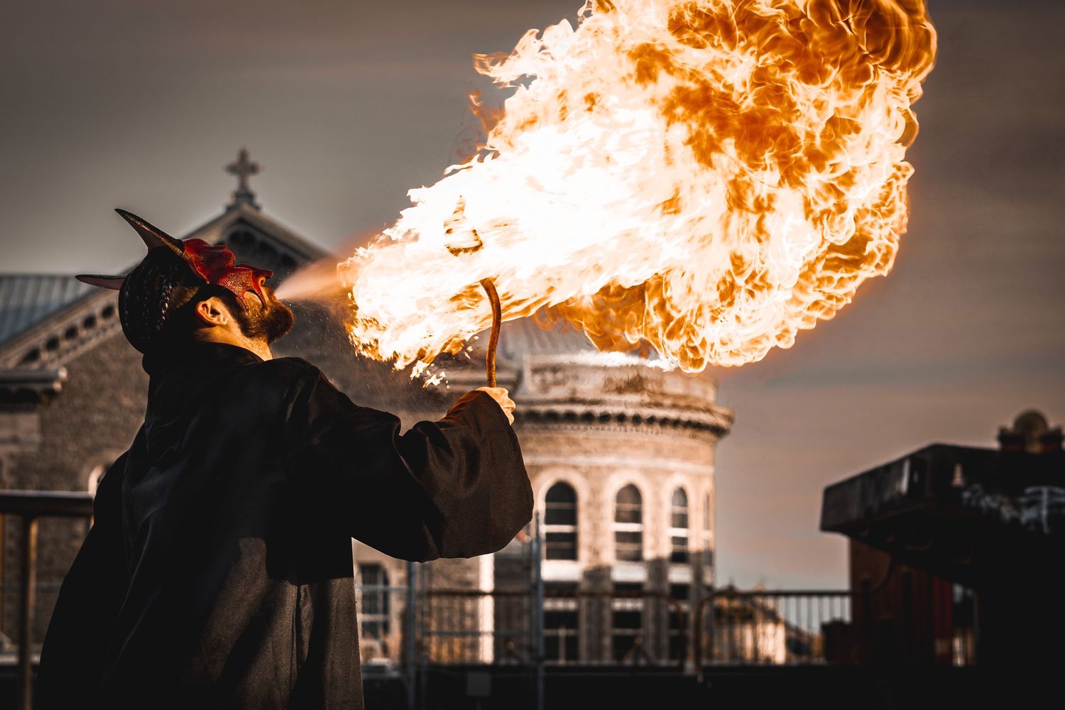 A fire breather performs on the street at dusk with a dramatic burst of flames against an ornate building backdrop.