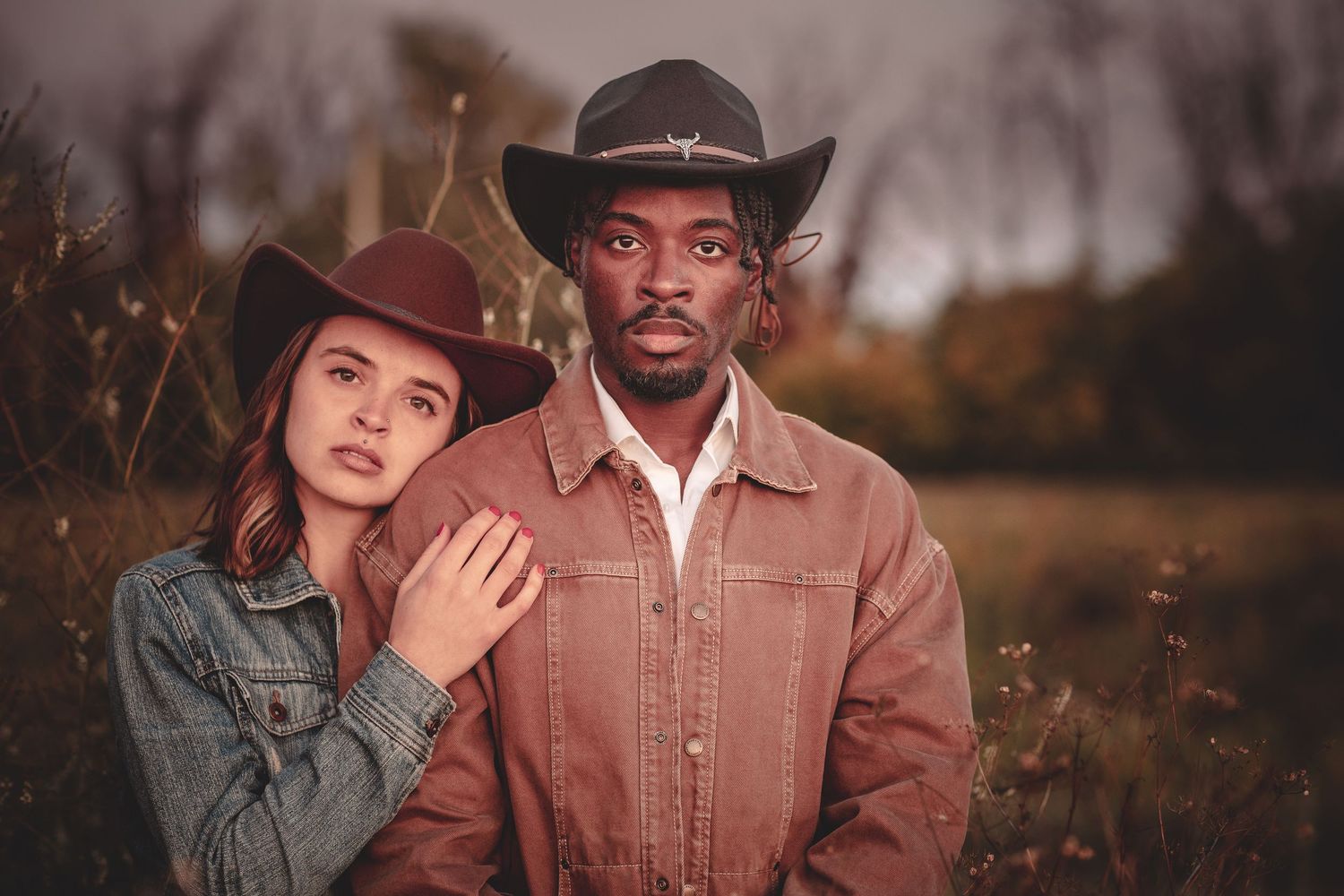 A romantic couple in western attire embraces during a golden hour photoshoot in a rustic outdoor setting.