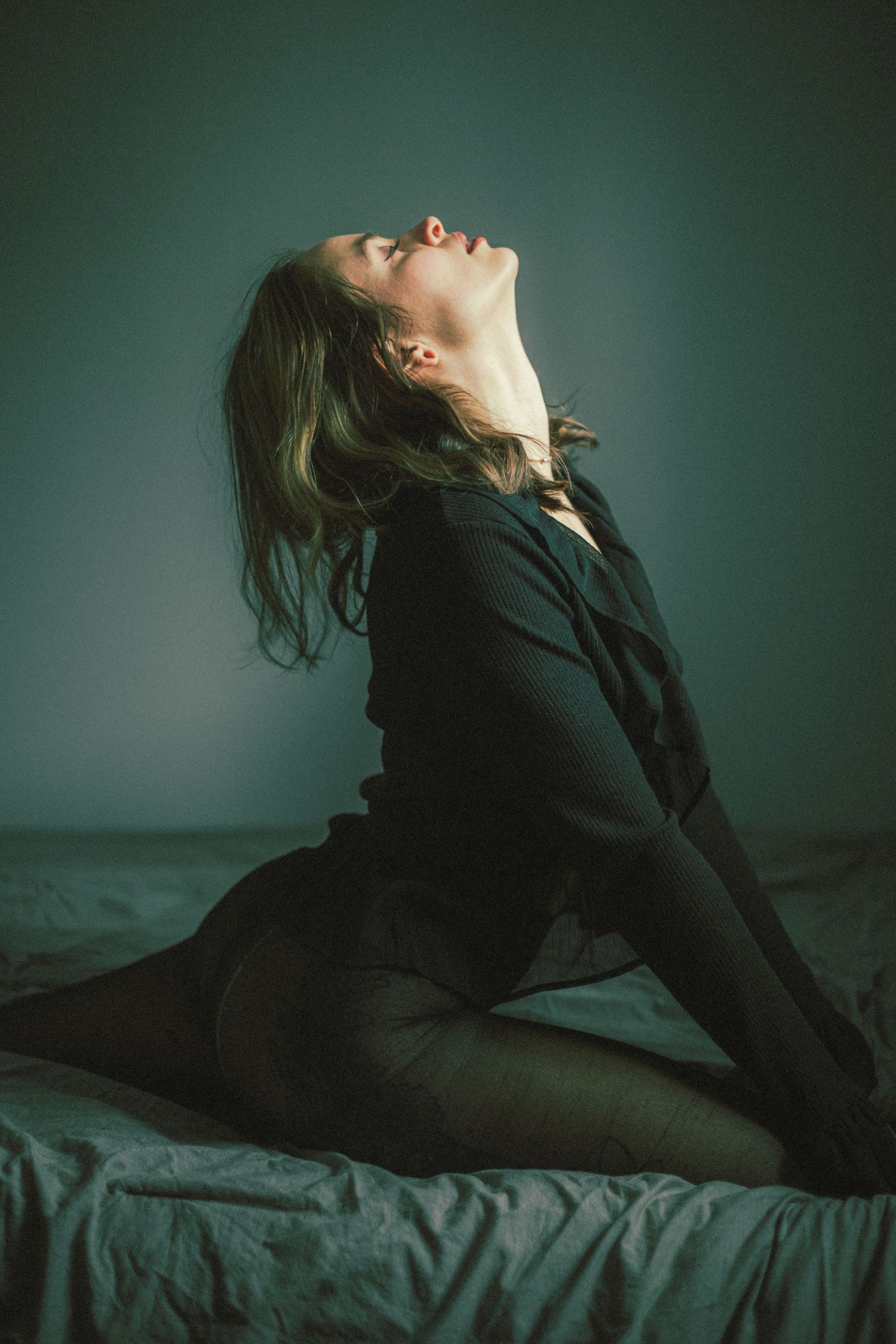 Artistic boudoir photo of person reclining on bed in dark clothing against teal wall.