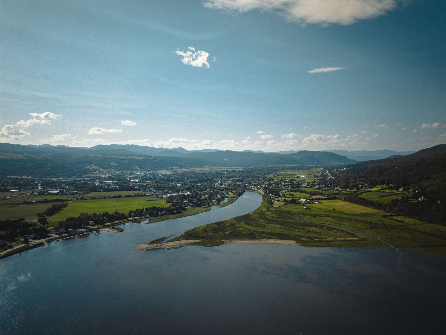 Aerial view of a winding river flowing through a green valley with mountains and blue sky in the background.