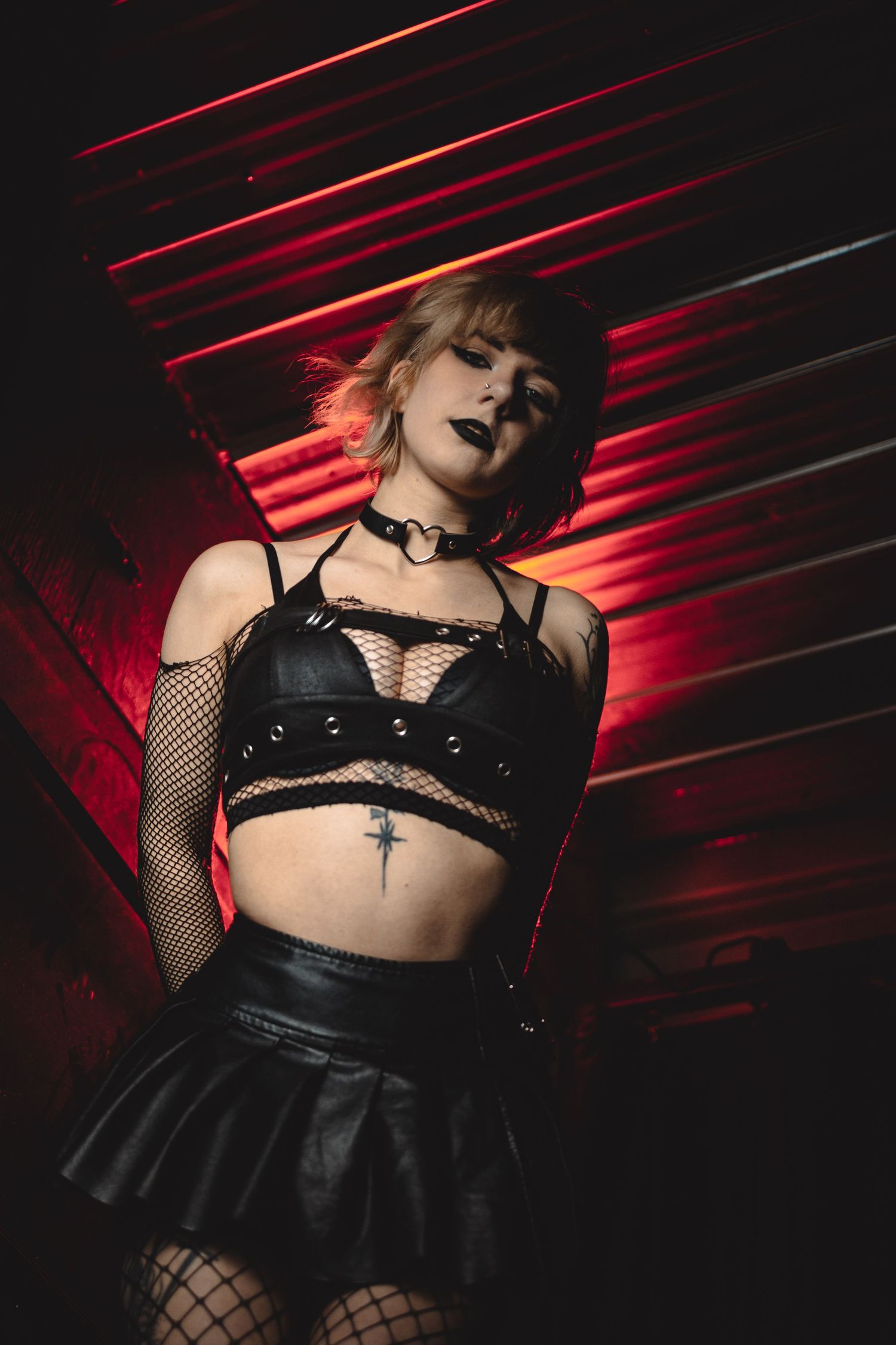 Gothic fashion model in black leather and fishnet against dramatic red lighting creates dark aesthetic atmosphere.