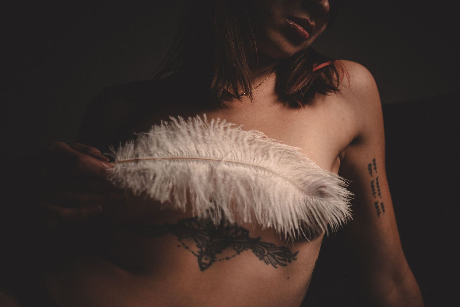 Artistic boudoir photo with white feather and tattoo detail in low-key lighting.