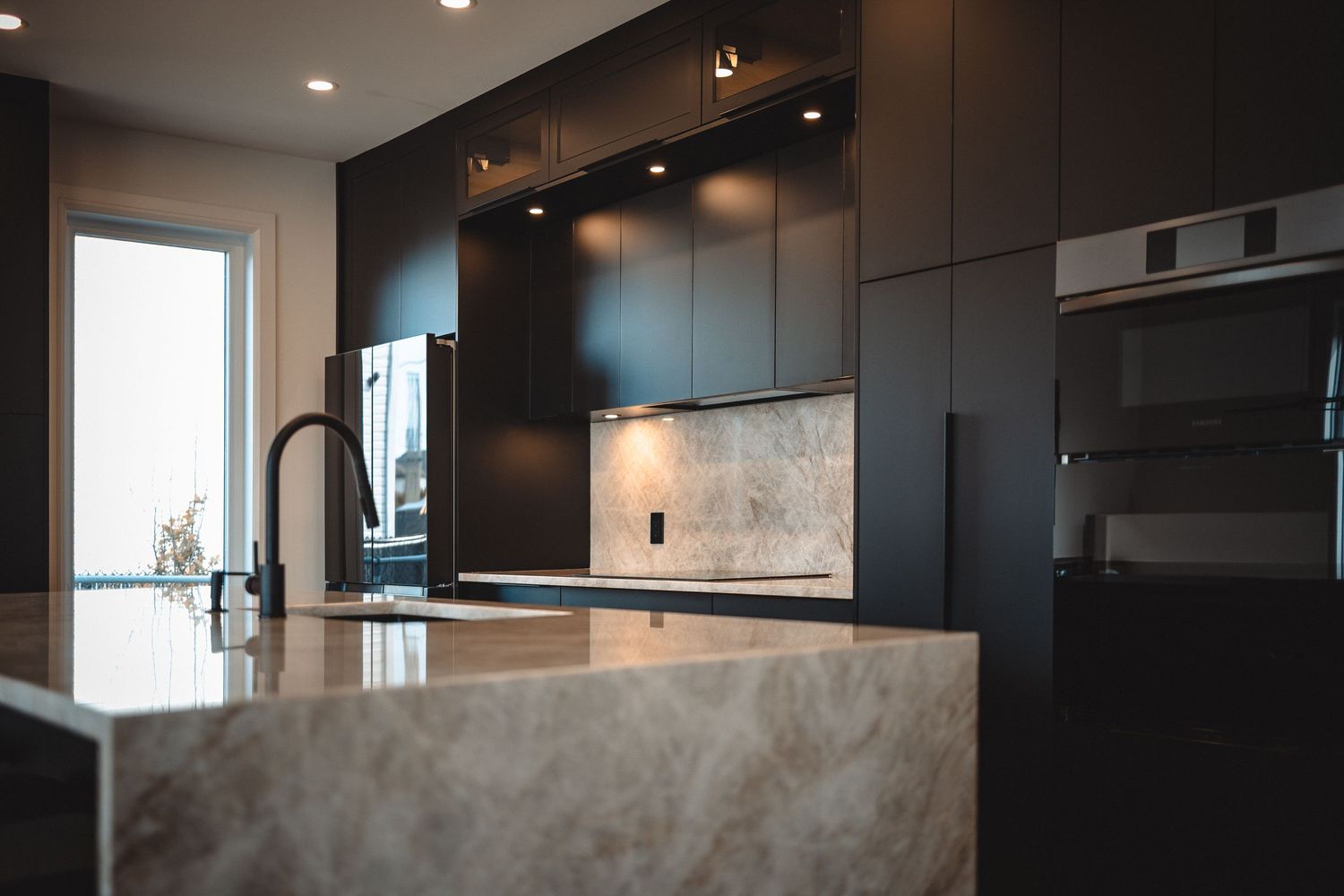 Contemporary kitchen featuring matte black cabinets and marble backsplash with dramatic lighting effects.