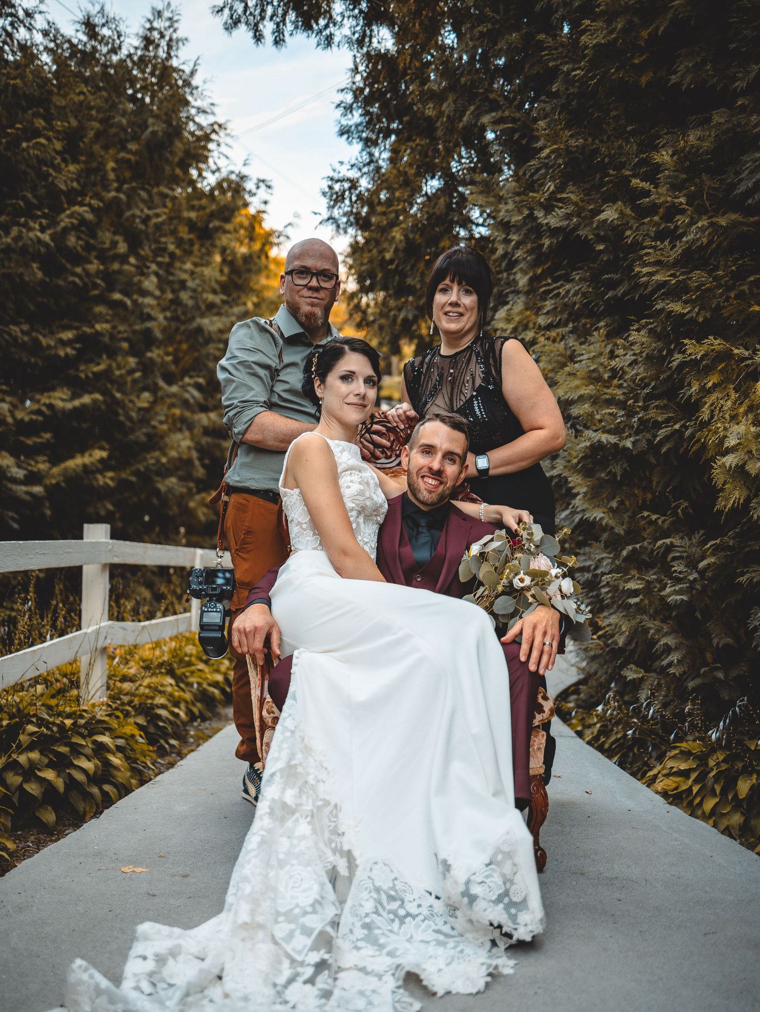 A couple in wedding attire poses playfully on a path surrounded by trees during an autumn day.