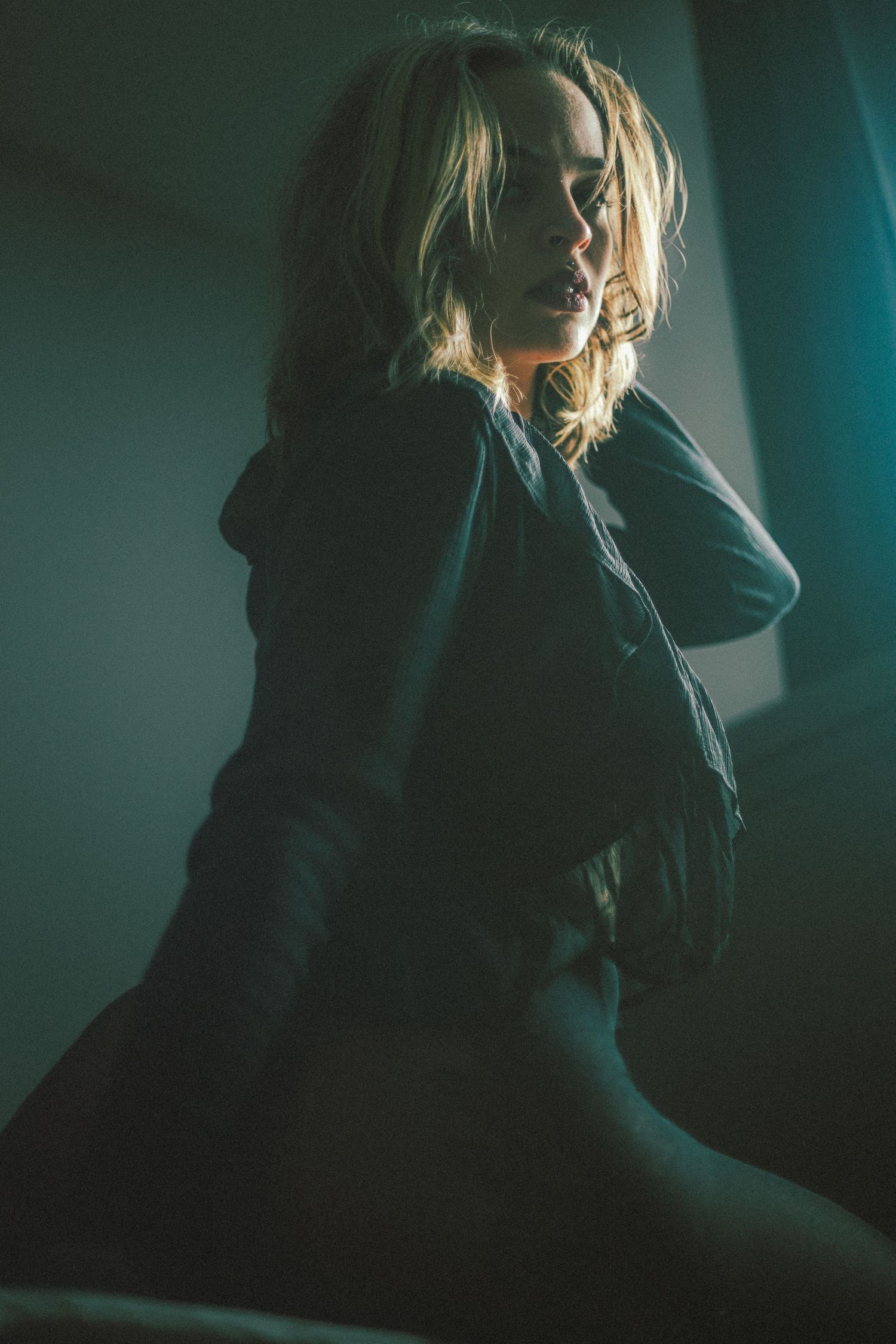 Moody portrait in dark tones with figure seated against teal background.