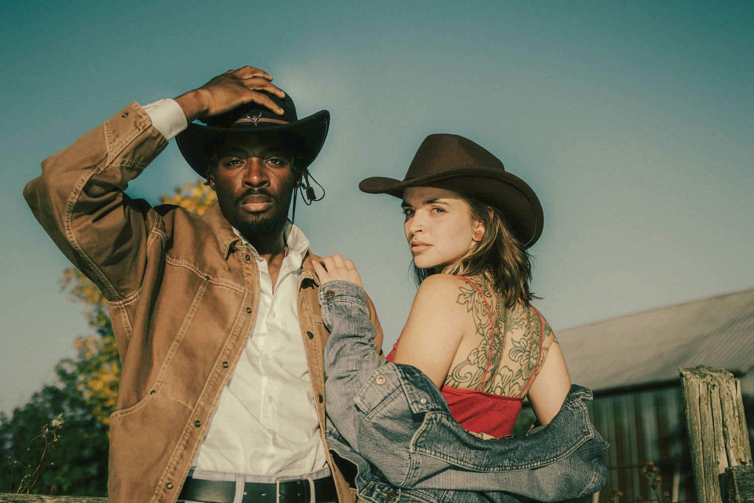 Two people in western cowboy attire pose together against a rustic outdoor backdrop during golden hour.