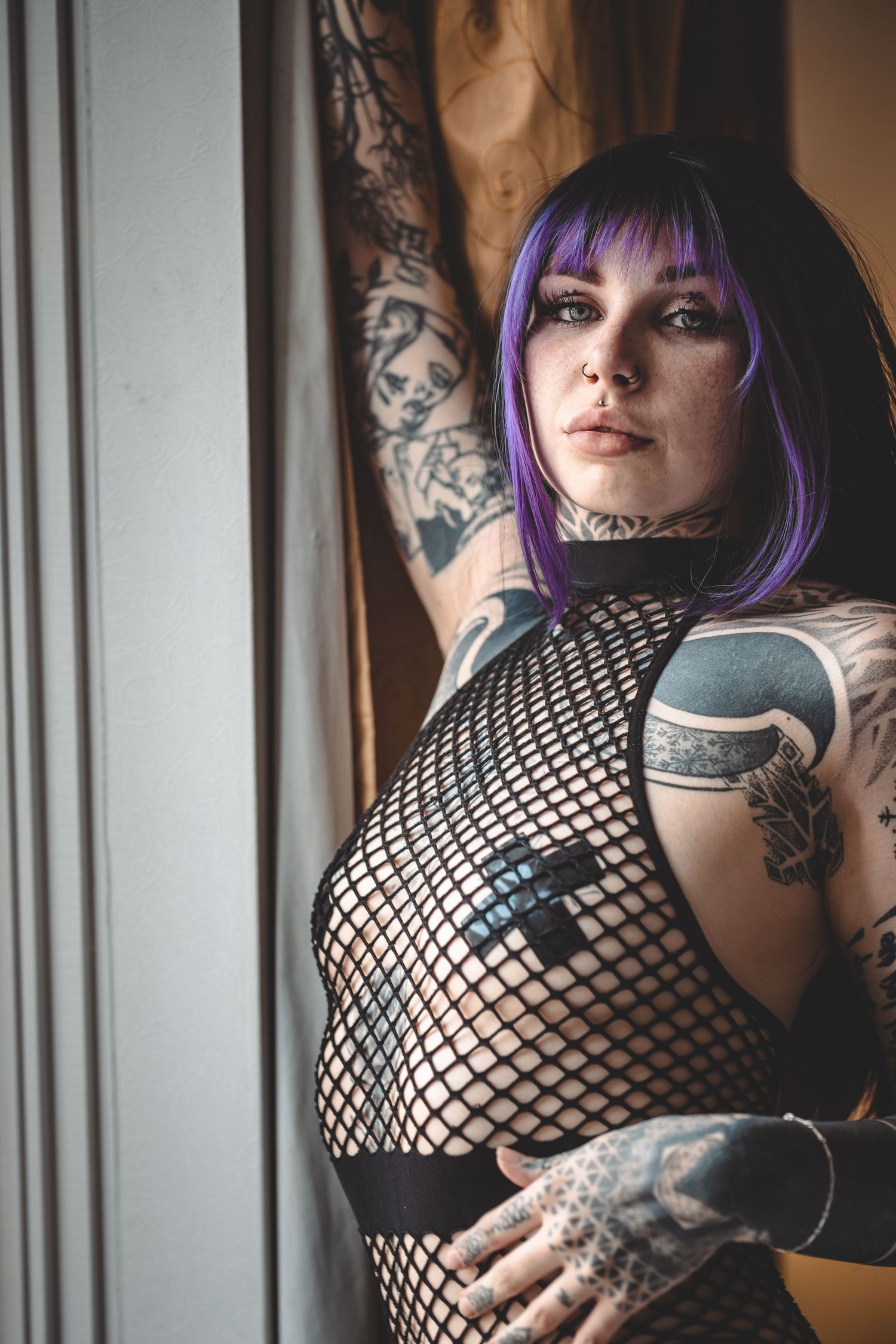 Alternative model wearing fishnet top with purple hair and tattoos in moody indoor lighting.
