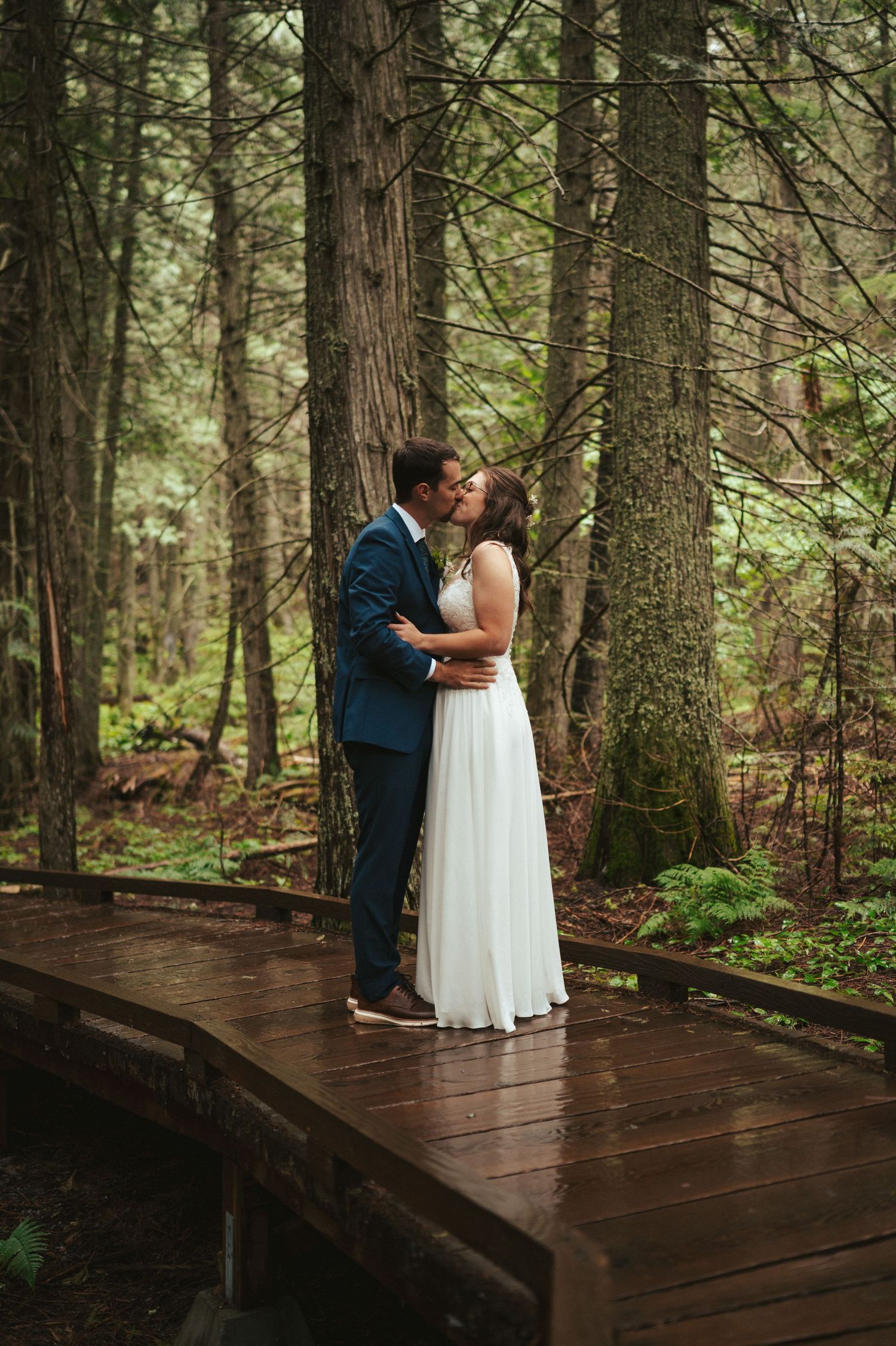 Romantic wedding couple embraces on a wooden deck surrounded by towering evergreen forest.