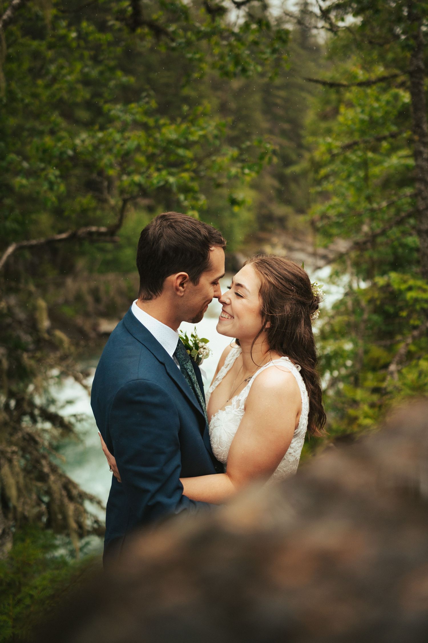 Romantic moment between newlyweds against scenic forest and water backdrop.