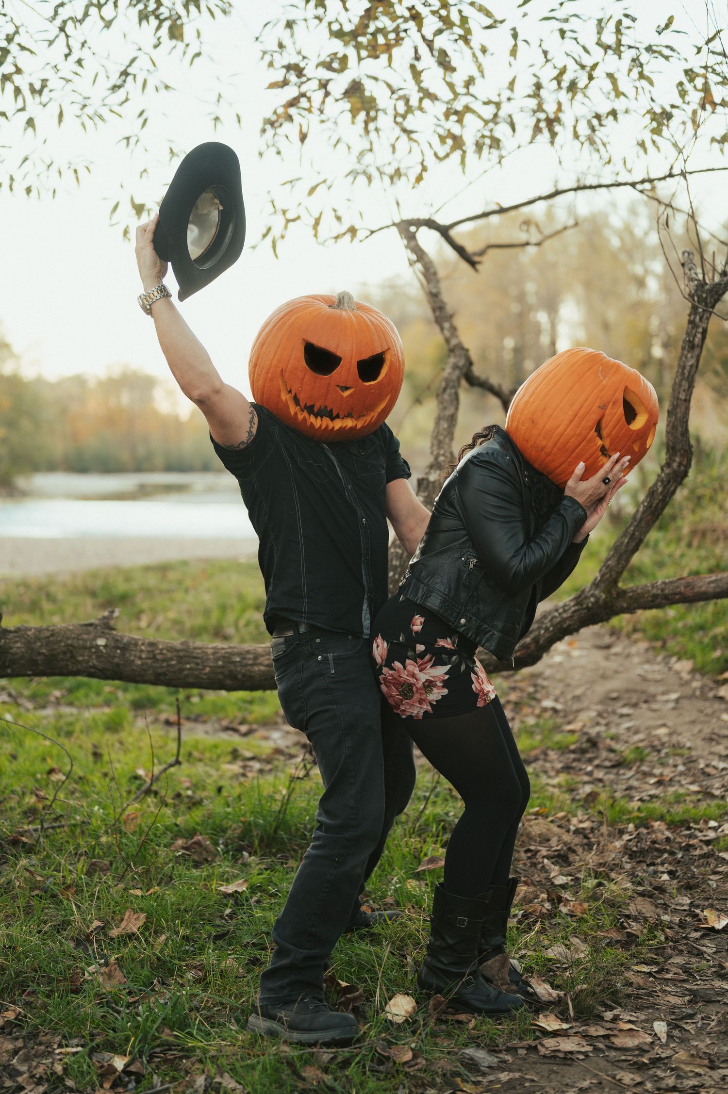 A playful Halloween couple in black outfits wearing jack-o-lantern masks poses outdoors in autumn by a lake.