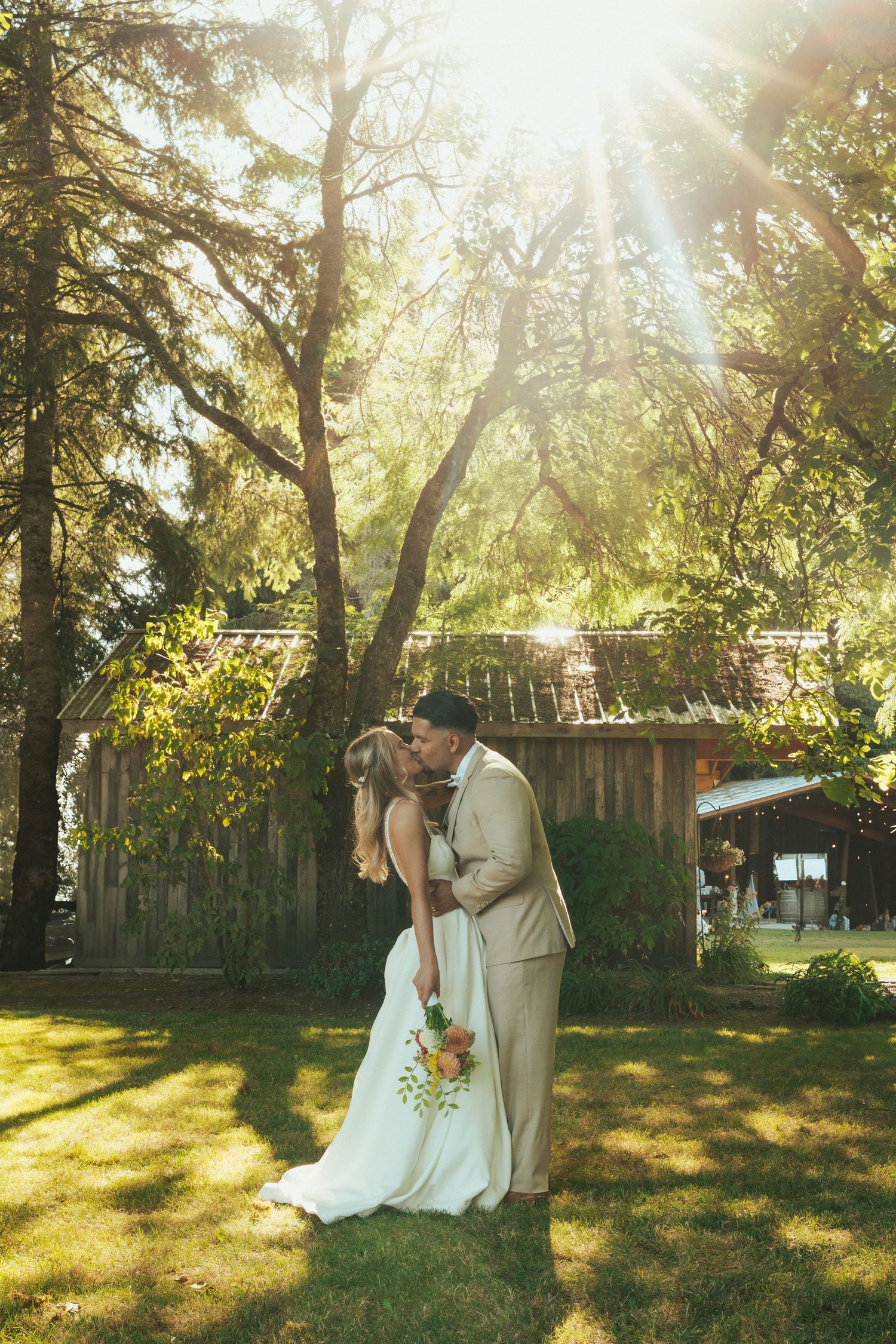 Newlyweds share a romantic kiss in golden sunlight beside a rustic barn.