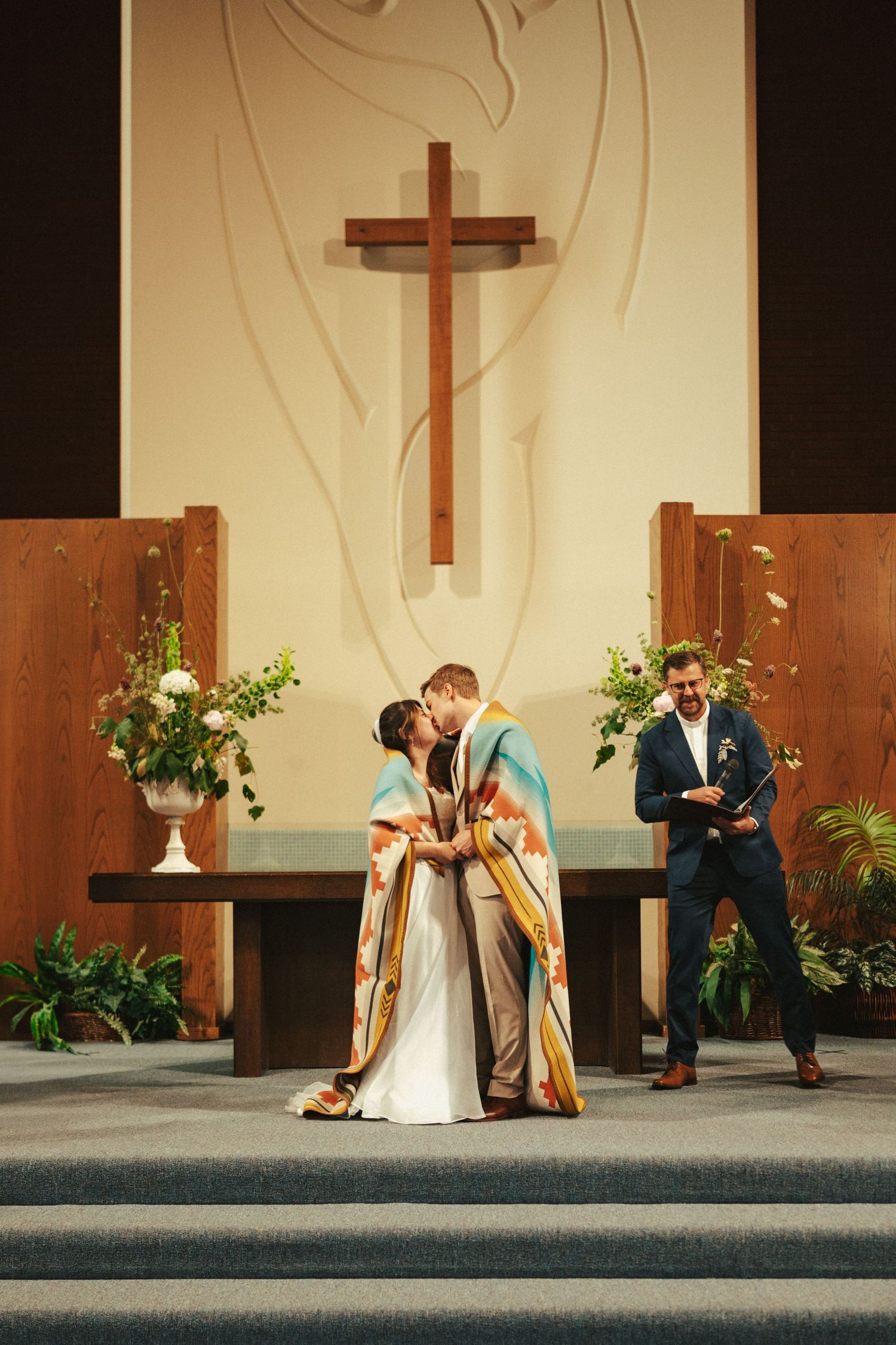 Wedding ceremony in church sanctuary with wooden cross and floral arrangements.