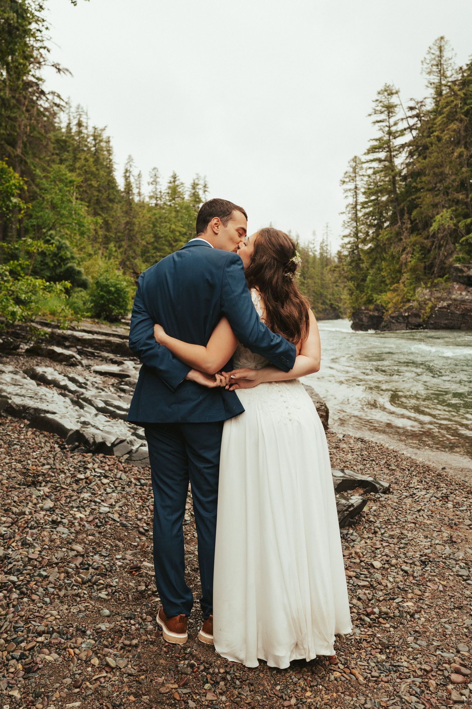 Newlyweds share tender moment on rocky beach with evergreen trees and mountains in background.
