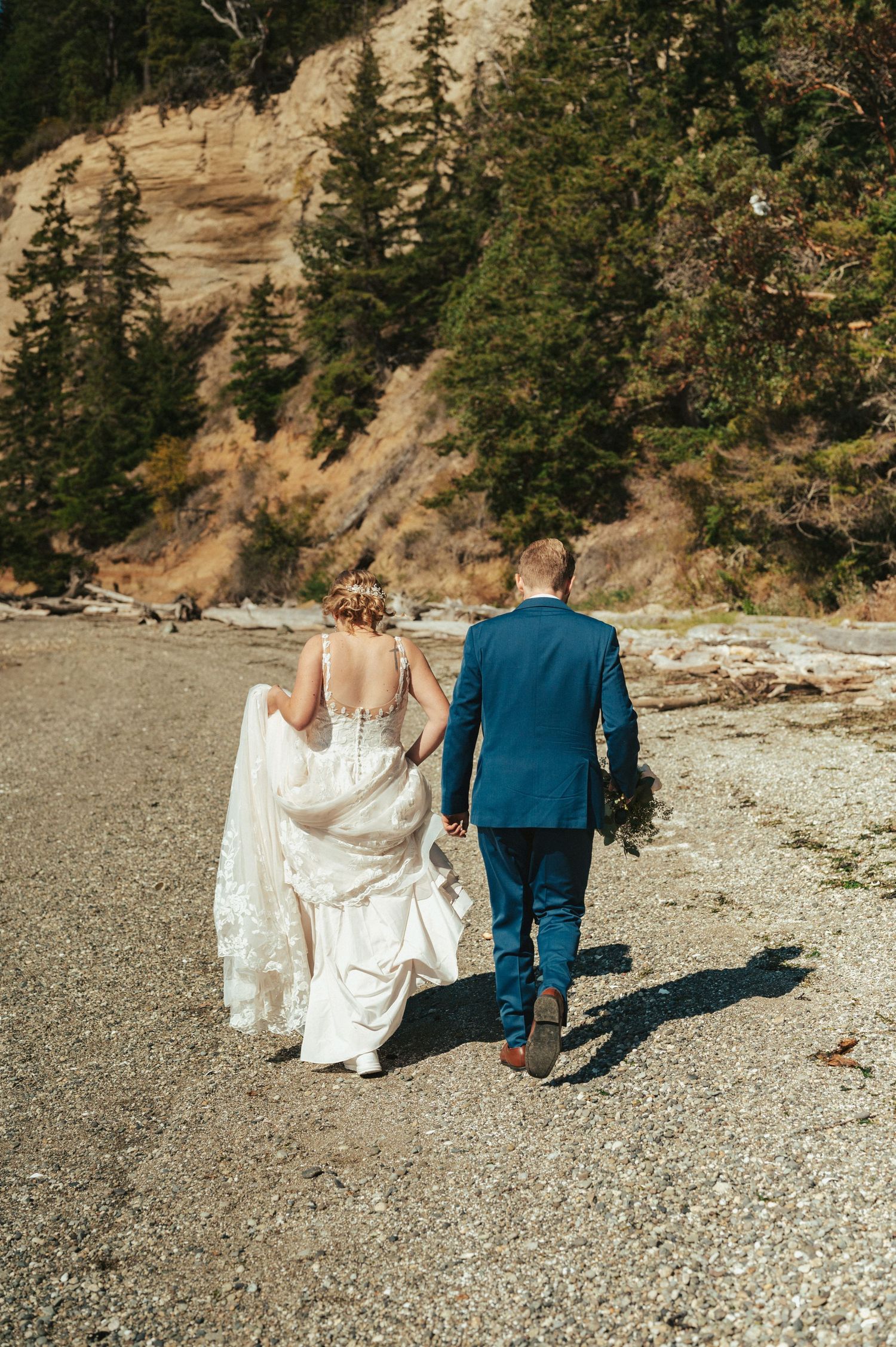 A couple walking on a beach path with dramatic cliffs and evergreen trees in the background.