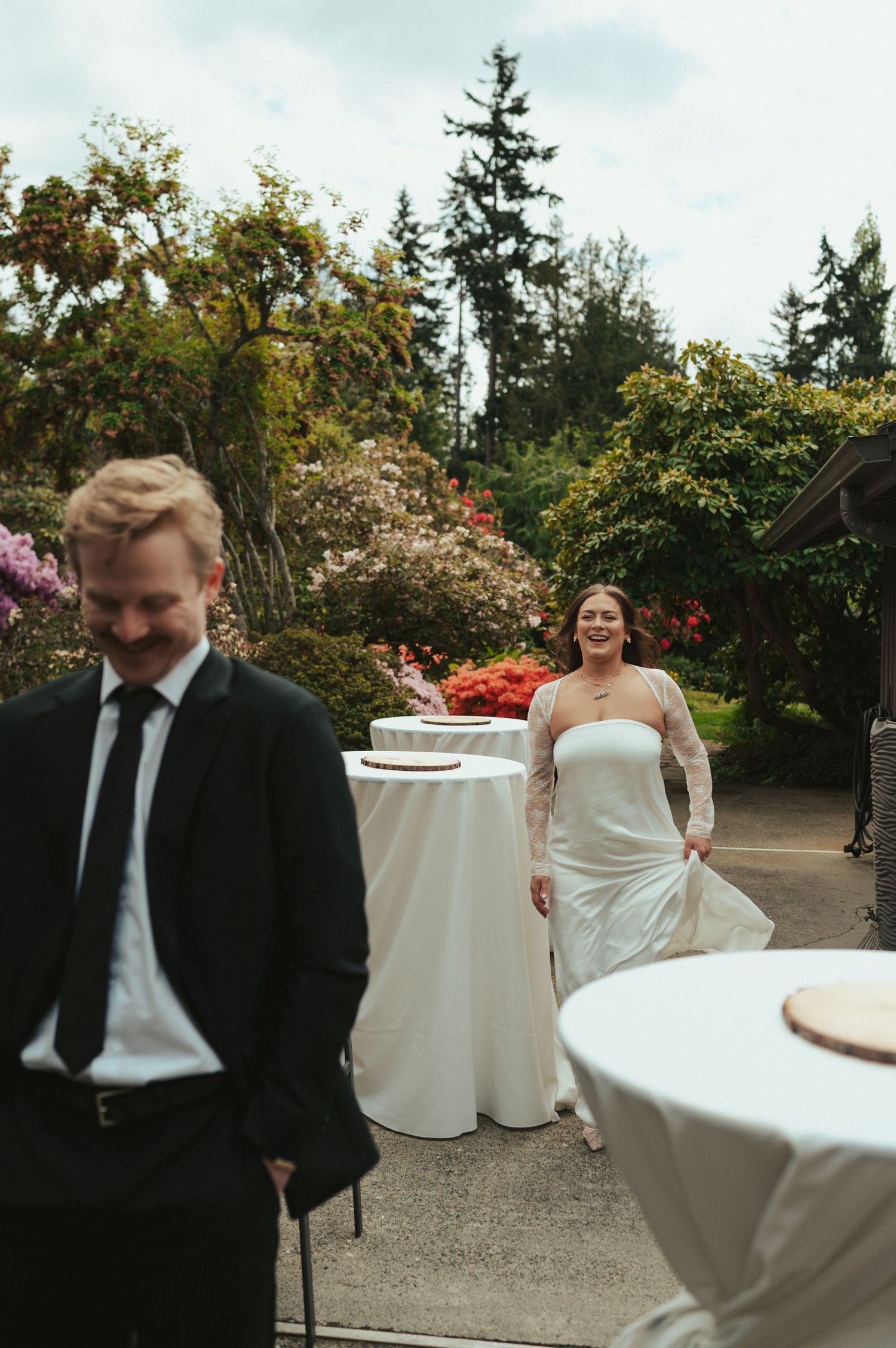 A joyous wedding reception moment with guests laughing around white-clothed tables in a garden setting.