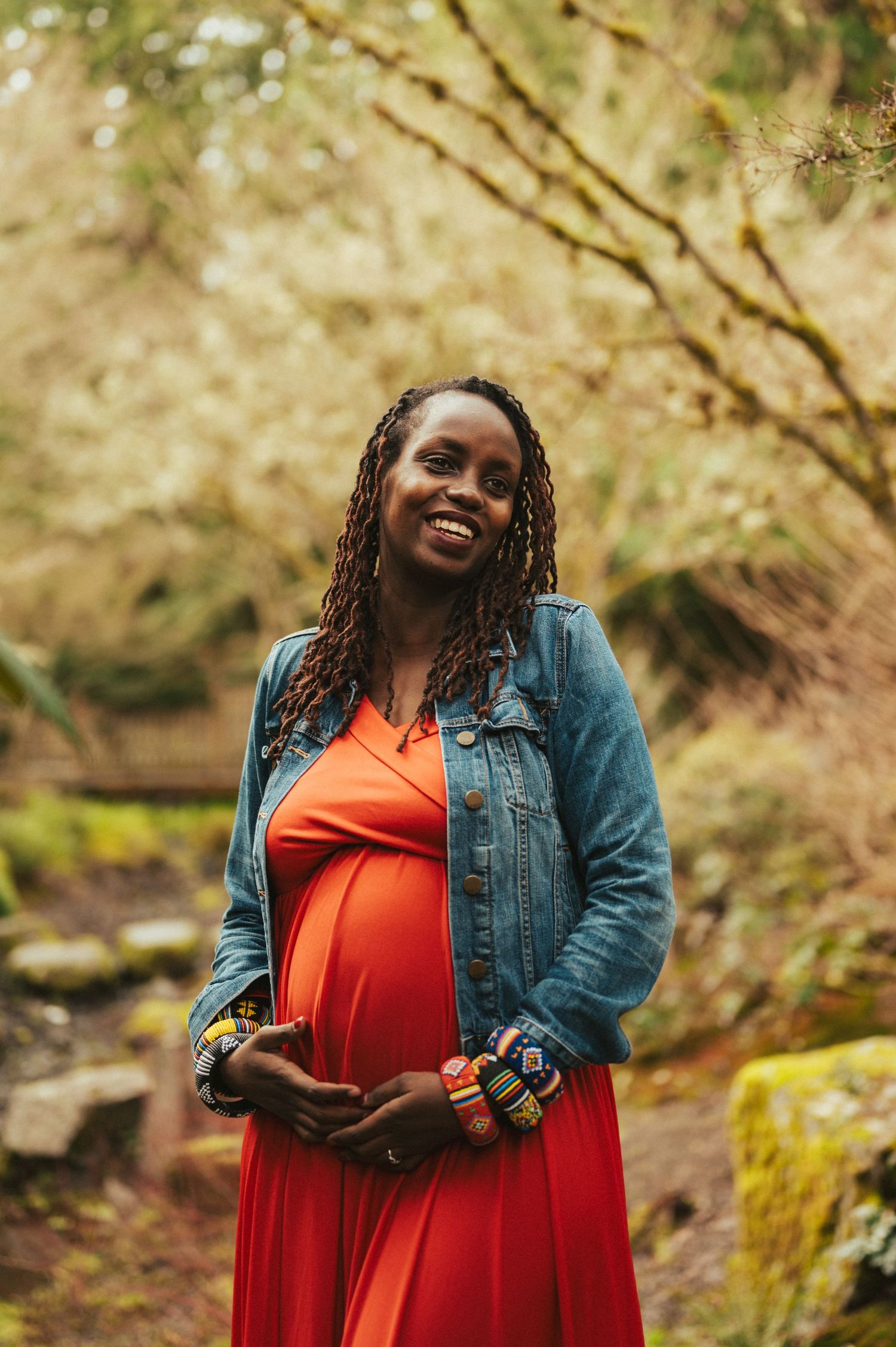 A person in a denim jacket and red dress smiles joyfully in a natural outdoor setting.