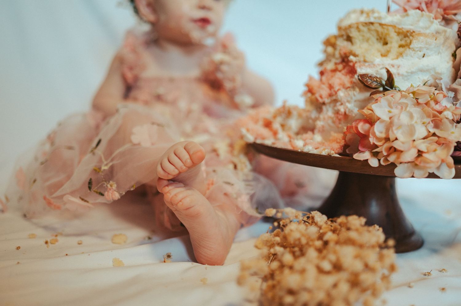 A baby in a pink dress sits near a smashed cake during a cake smash photo session.