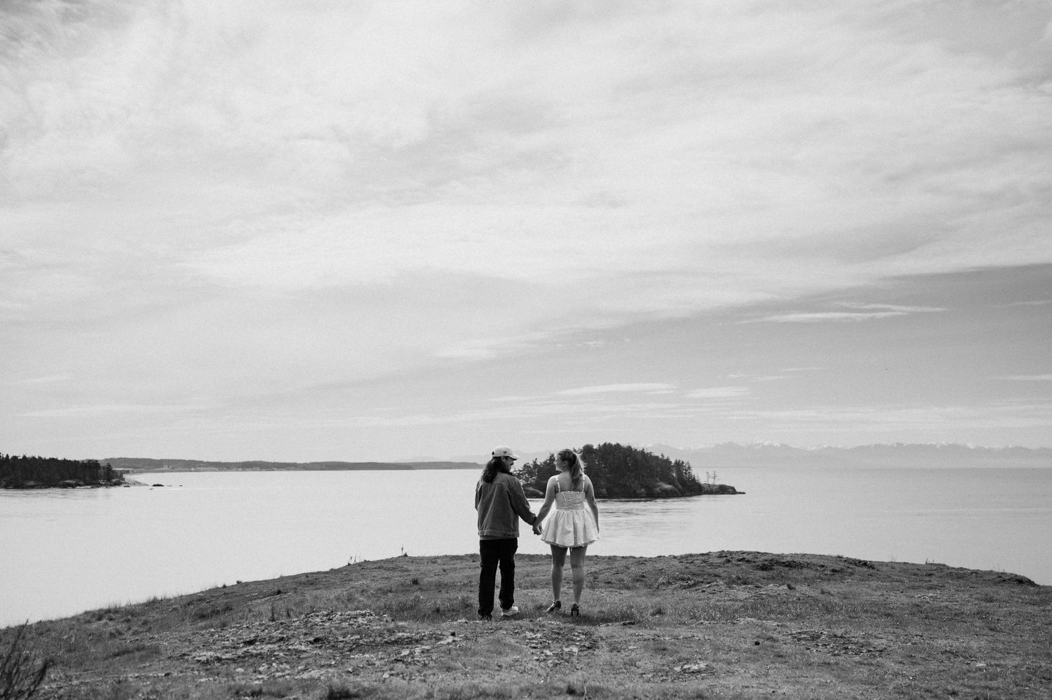 Two silhouettes stand together on a cliff overlooking a serene lake and distant islands.