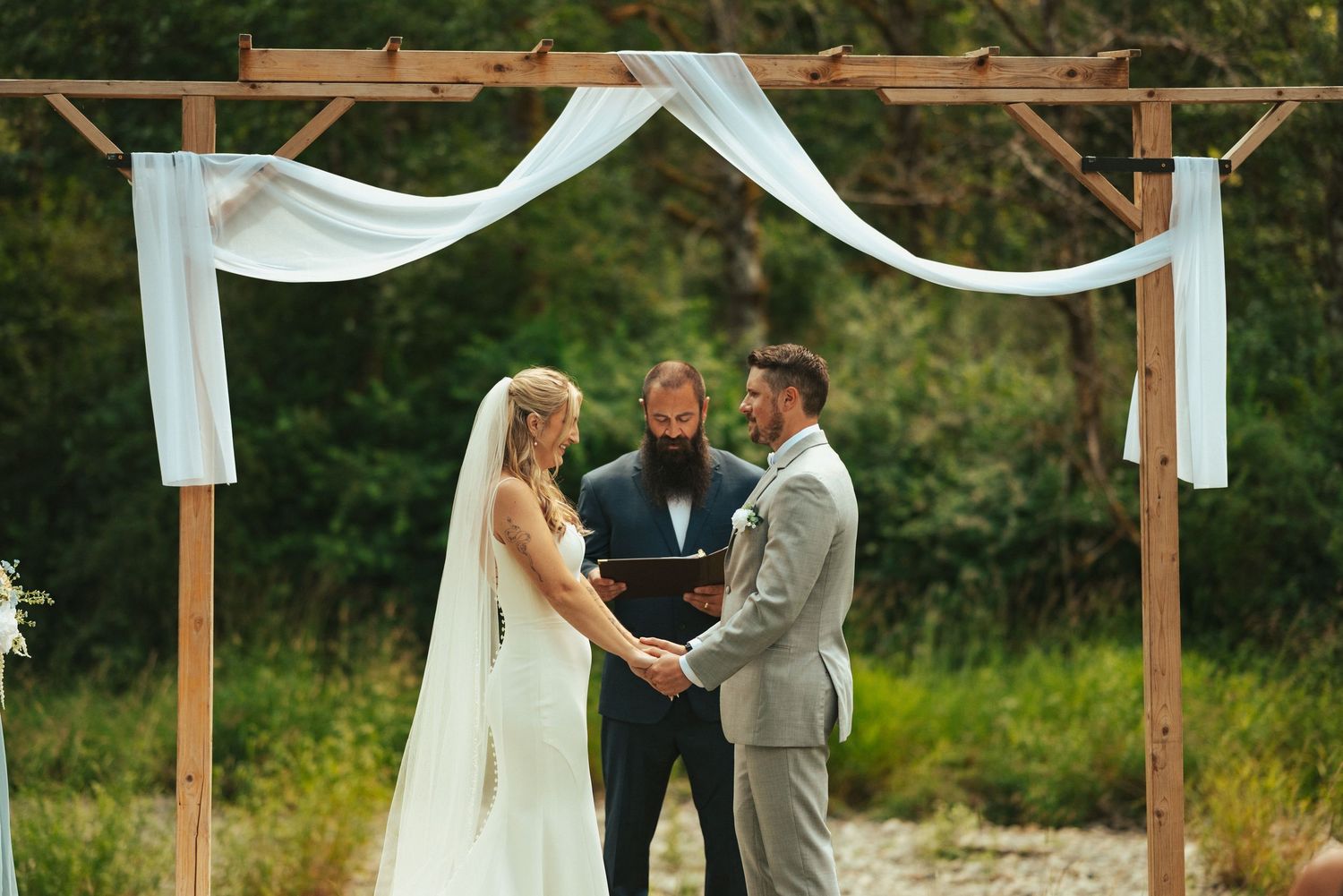 A wedding ceremony takes place under a rustic wooden arch decorated with flowing white fabric in an outdoor setting.