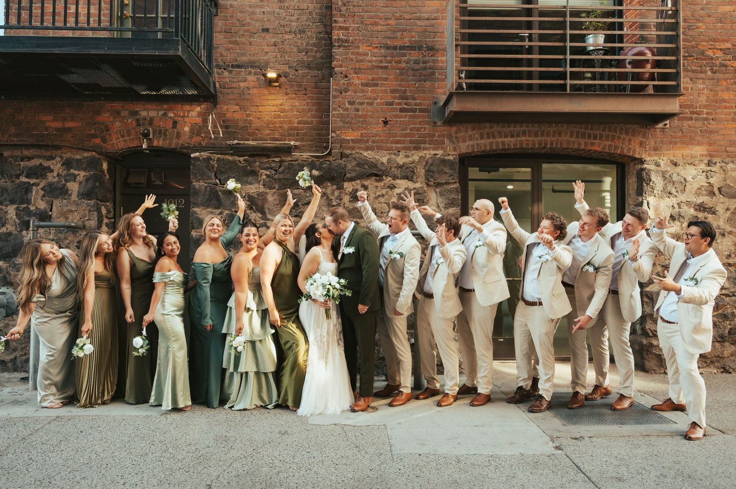 Wedding party members lined up against a rustic brick wall for group photos during an outdoor celebration.