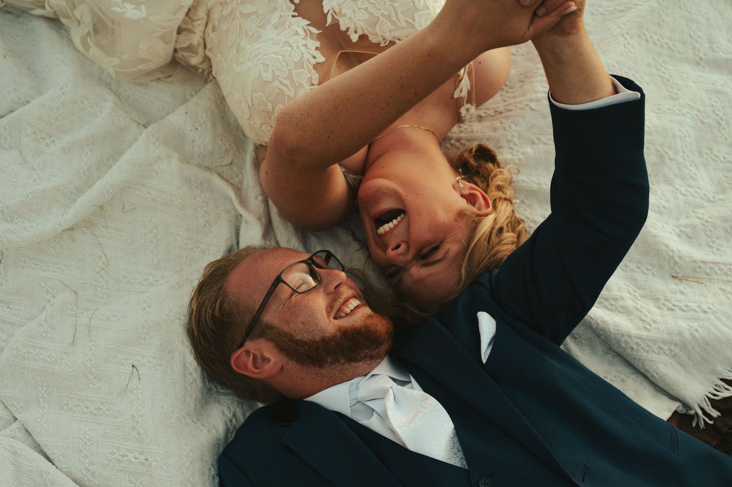 A joyful wedding couple sharing a candid laughing moment while laying down together.
