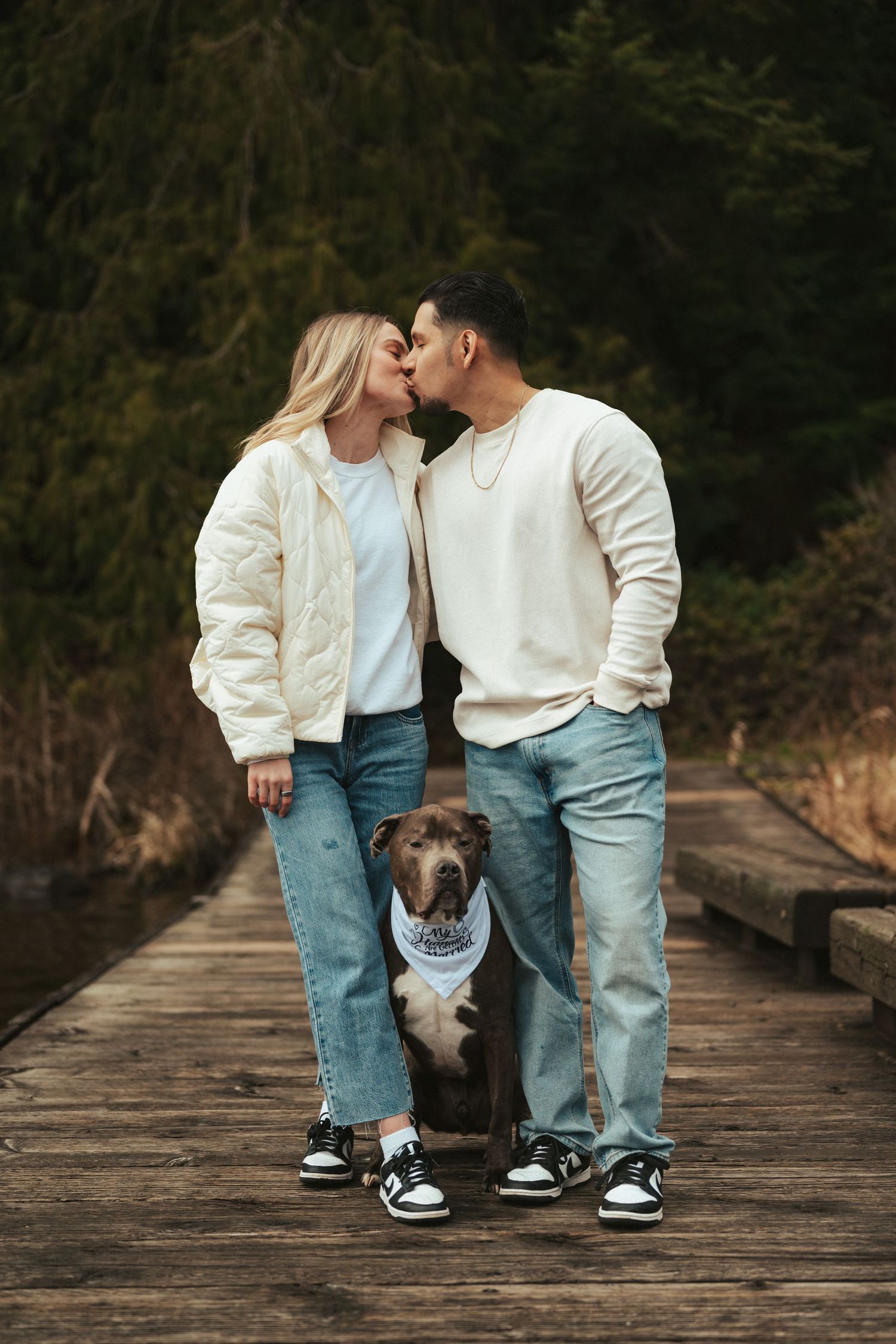 A couple shares a kiss on a wooden bridge with their dog between them.