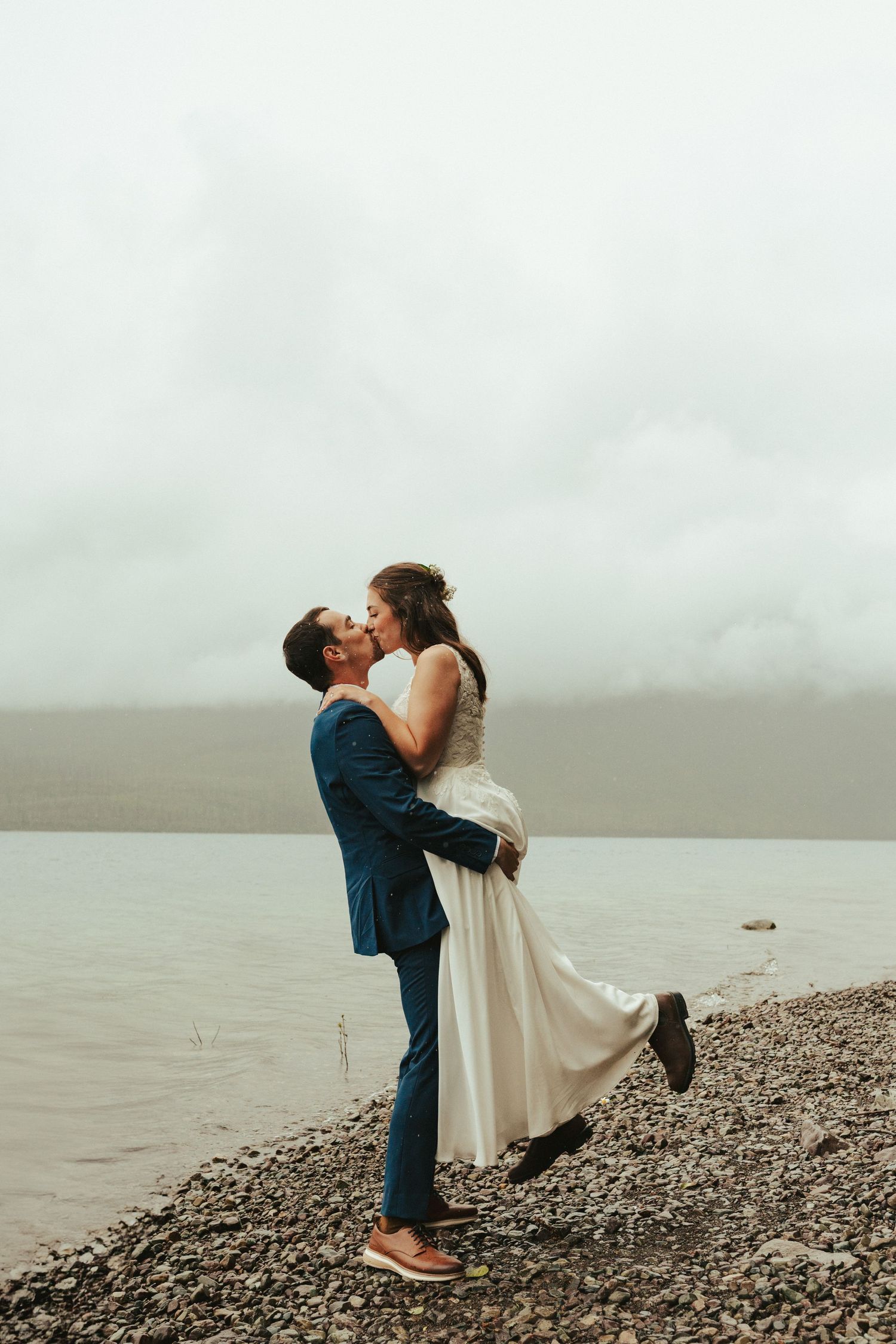 Wedding couple's romantic lift pose on pebble beach with misty water backdrop.