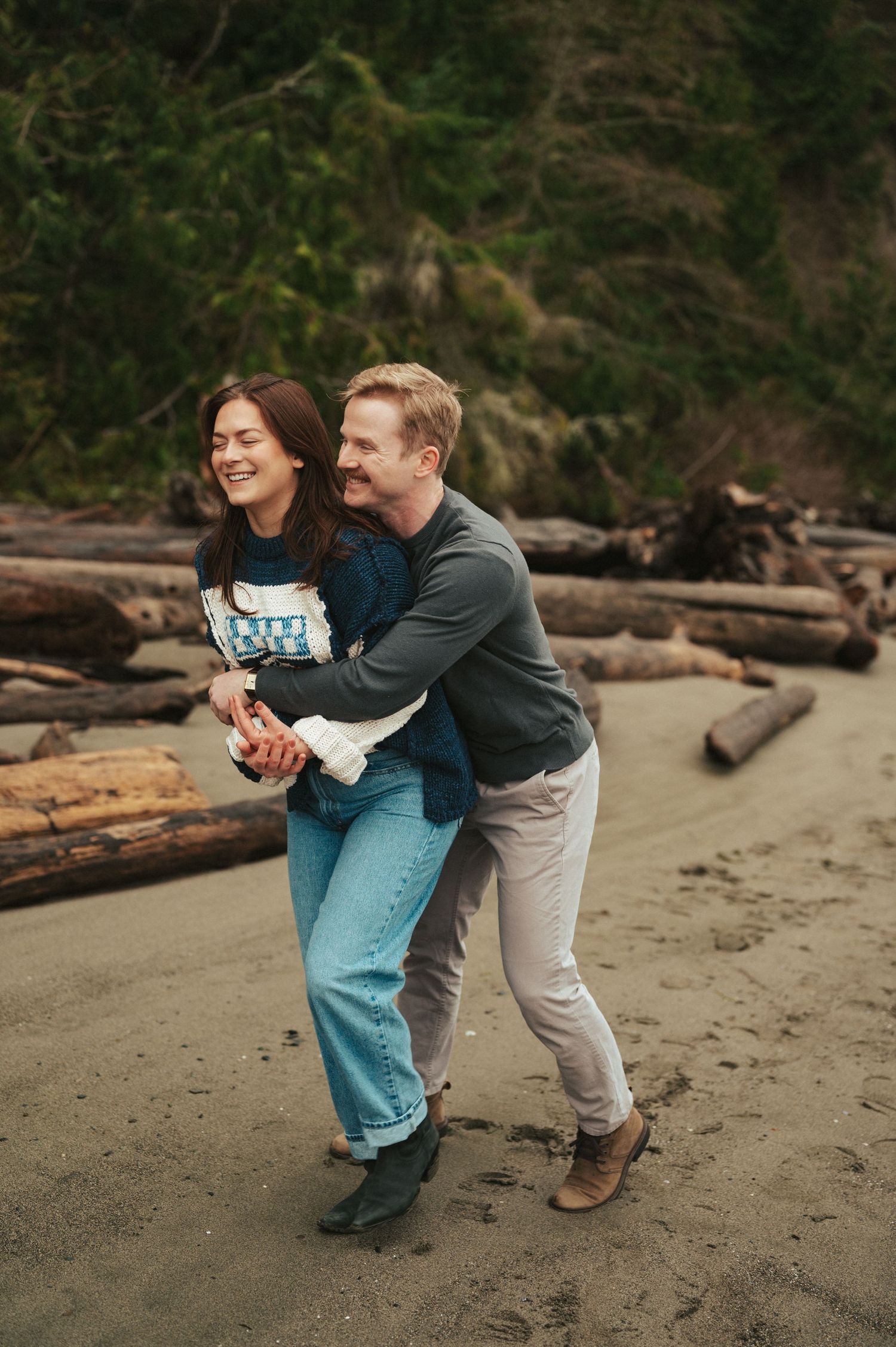 A joyful embrace on a driftwood-strewn beach with forest backdrop.