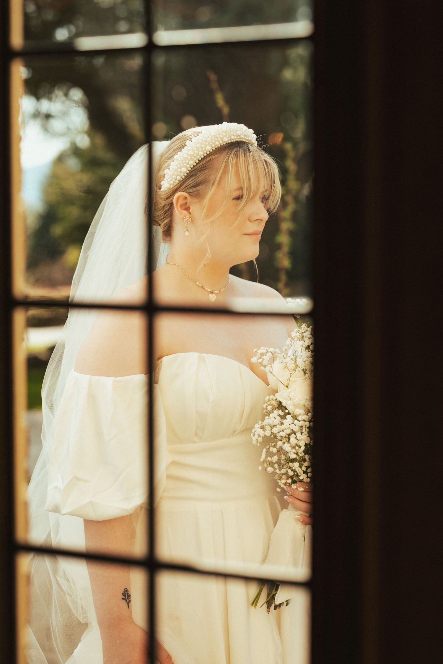 A bride wearing a white dress and veil is photographed through a window pane in warm sunlight.