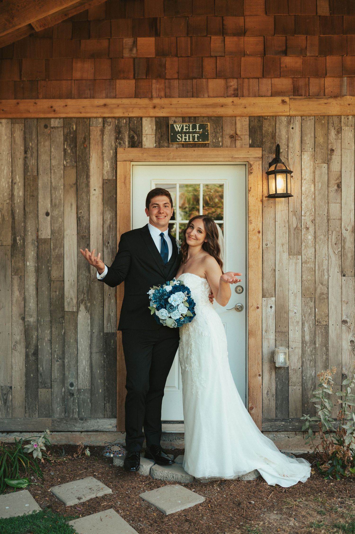 Newlyweds posing in front of rustic wooden cabin with blue bouquet.