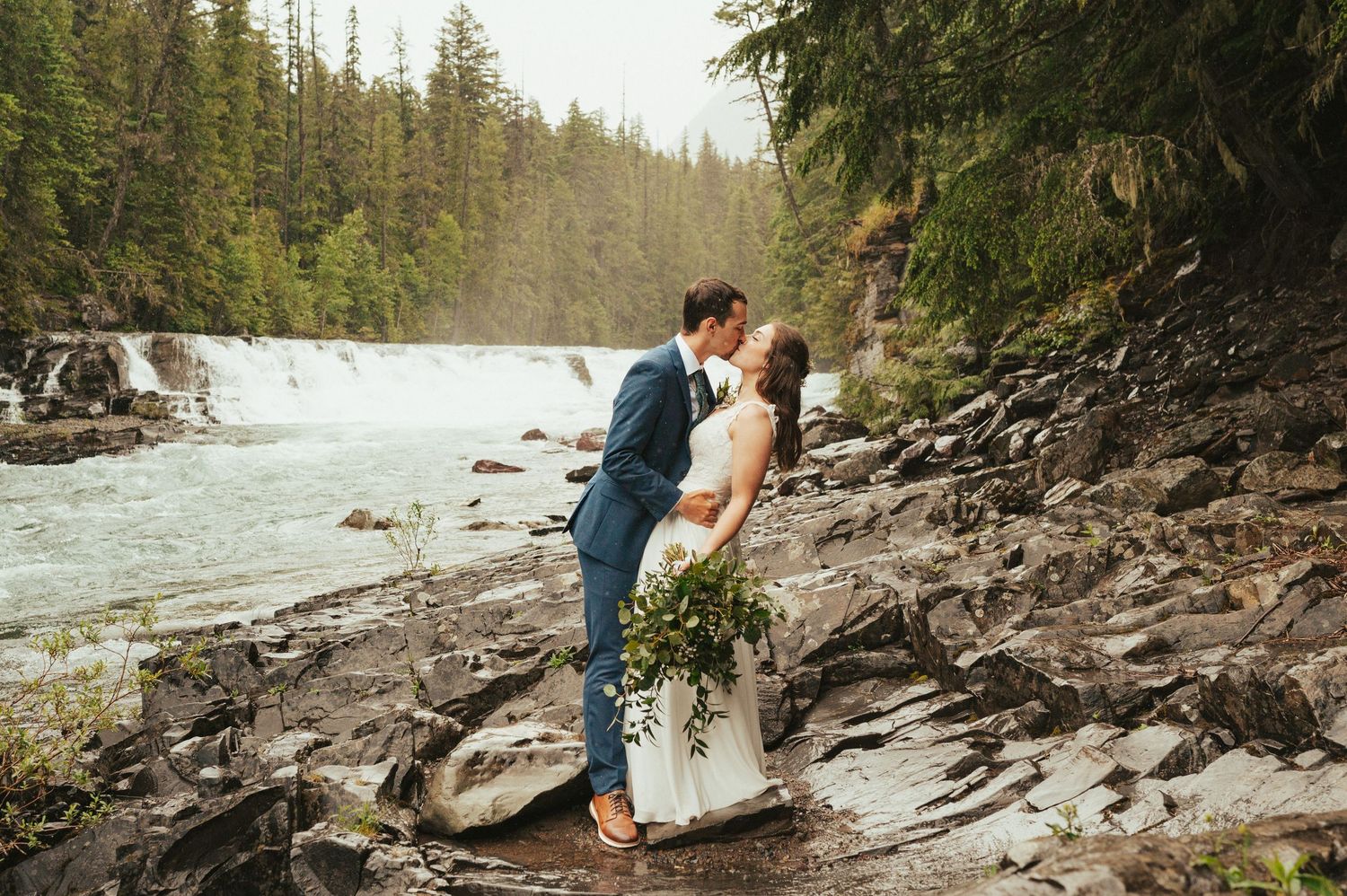A romantic wedding photo on rocky terrain with a rushing waterfall and forest backdrop.