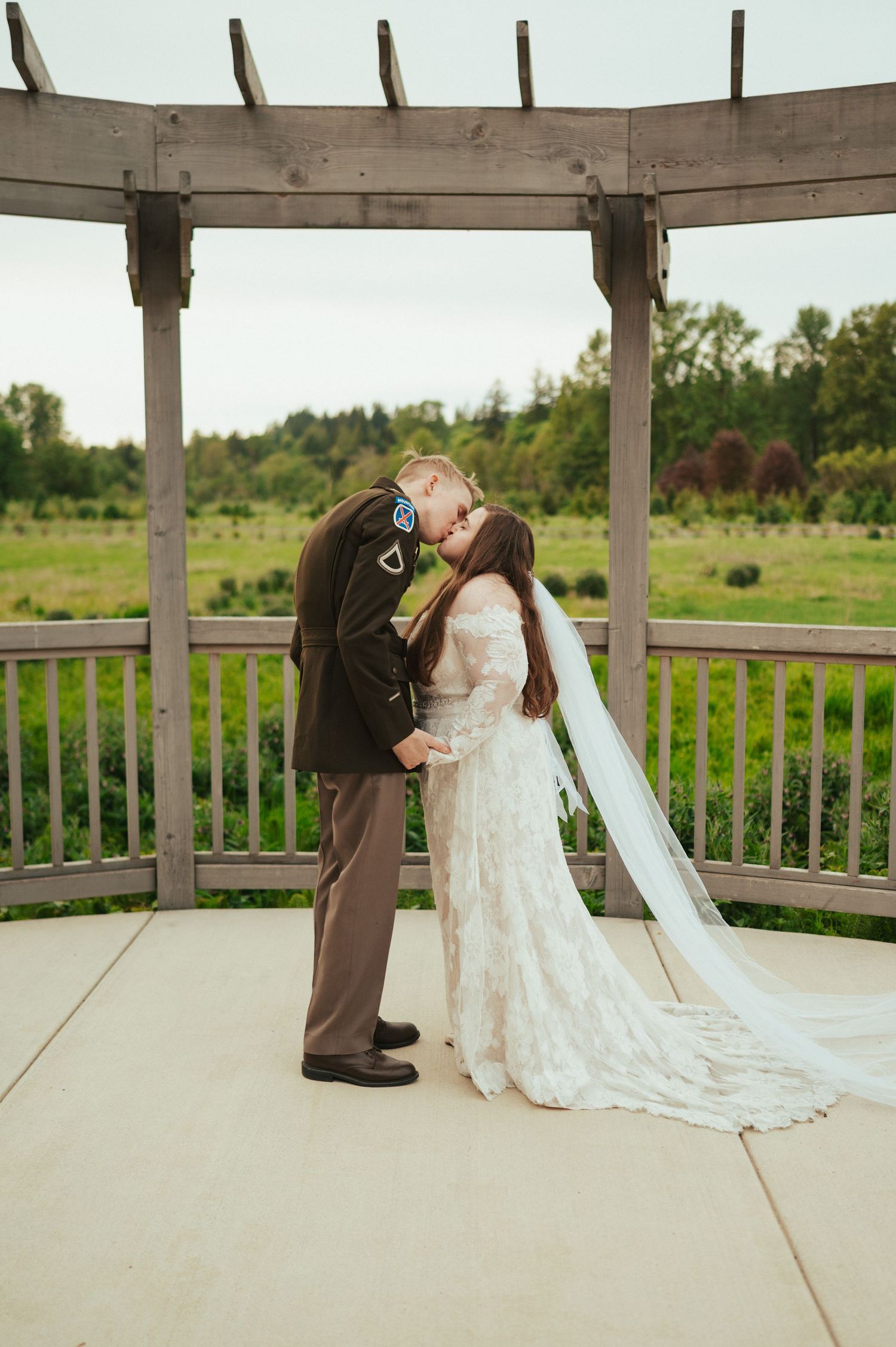 A romantic wedding moment captured under a wooden pergola in a garden setting with a long flowing white lace dress and veil.