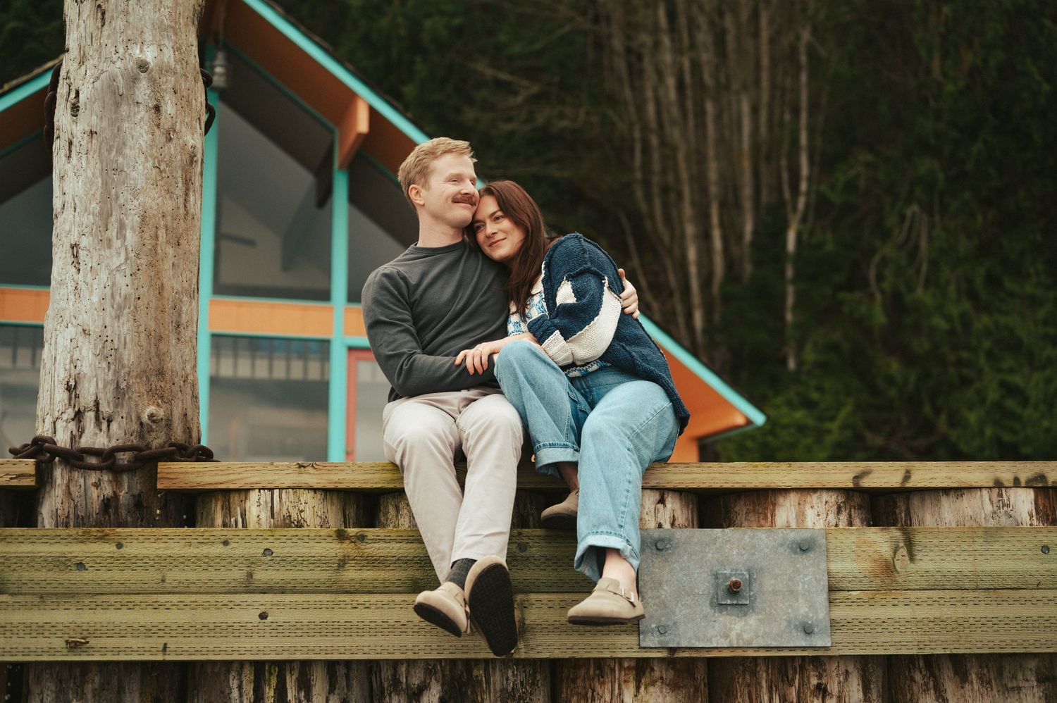 A couple sits with their dog on a wooden dock by a cabin in the woods.