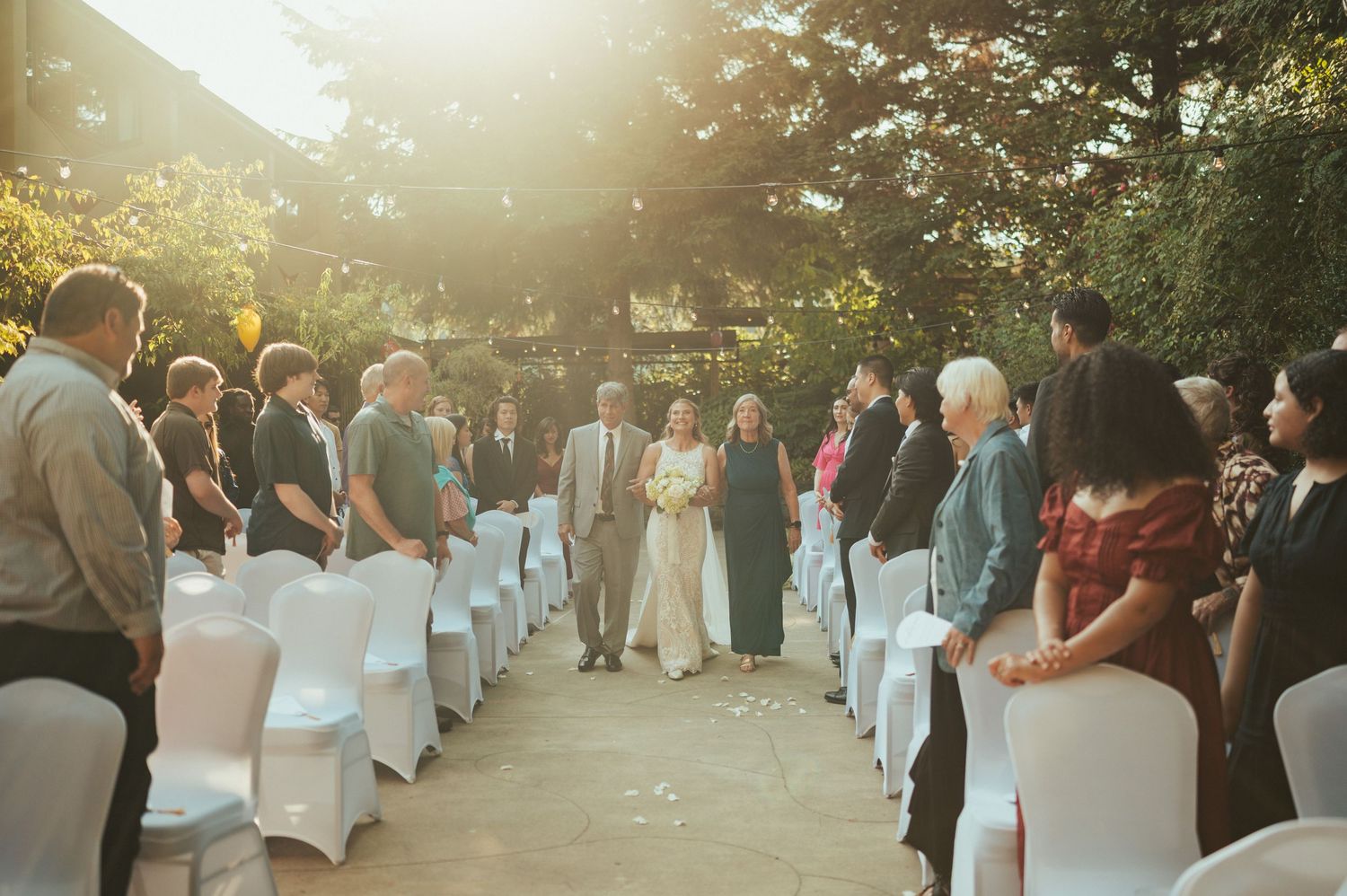 Wedding guests seated in white chairs at an outdoor ceremony during golden hour sunset.