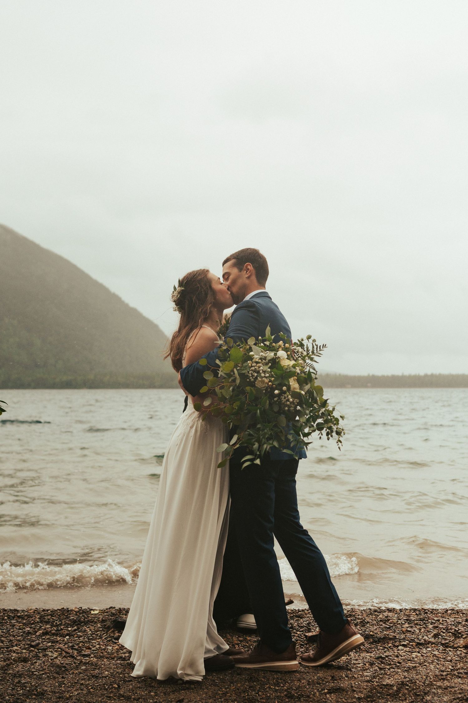 A couple shares a romantic kiss on a misty beach shore with mountains and water in the background.