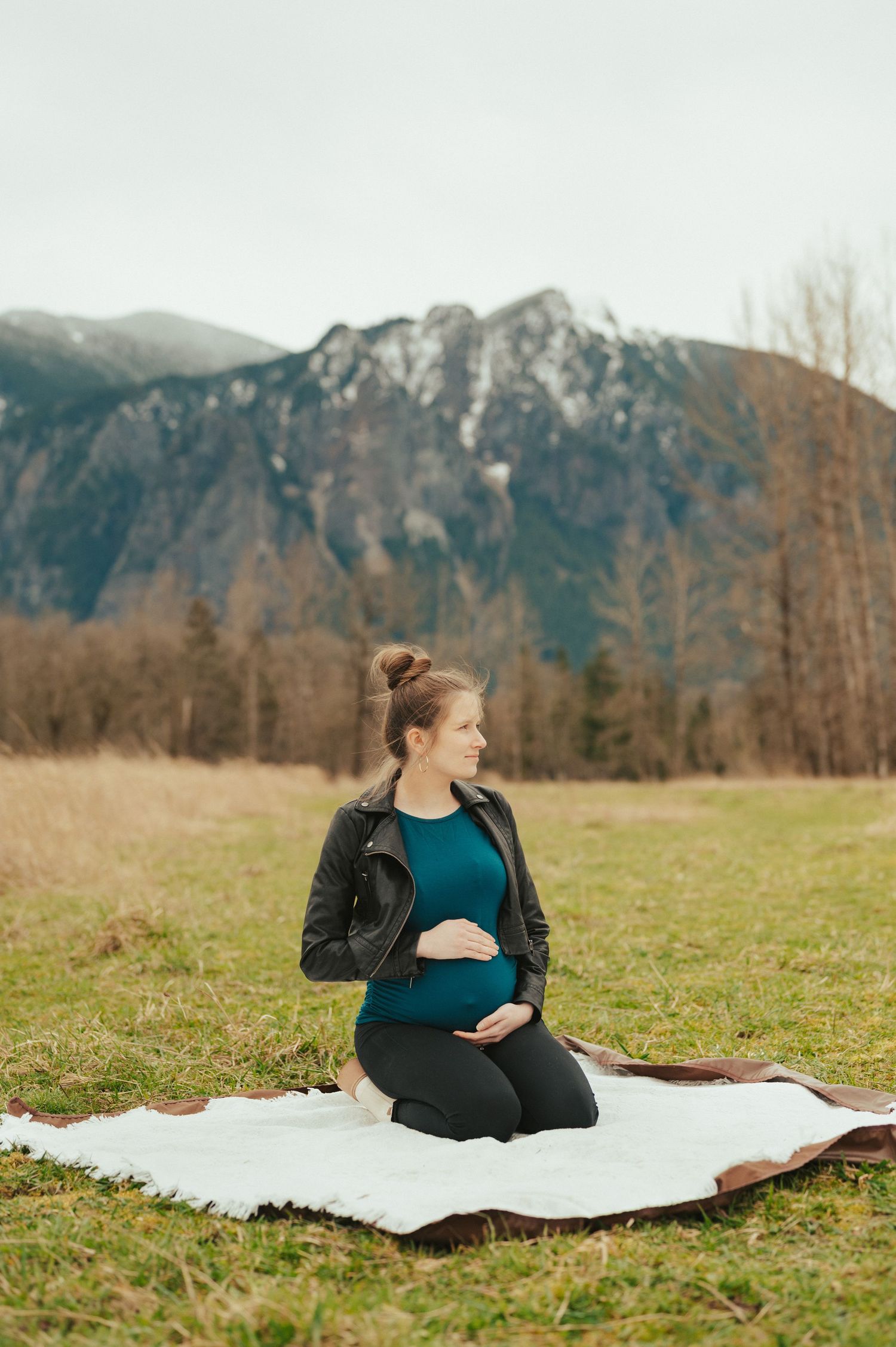 A maternity photoshoot with mountains in background showing someone resting on blanket in a grassy field.