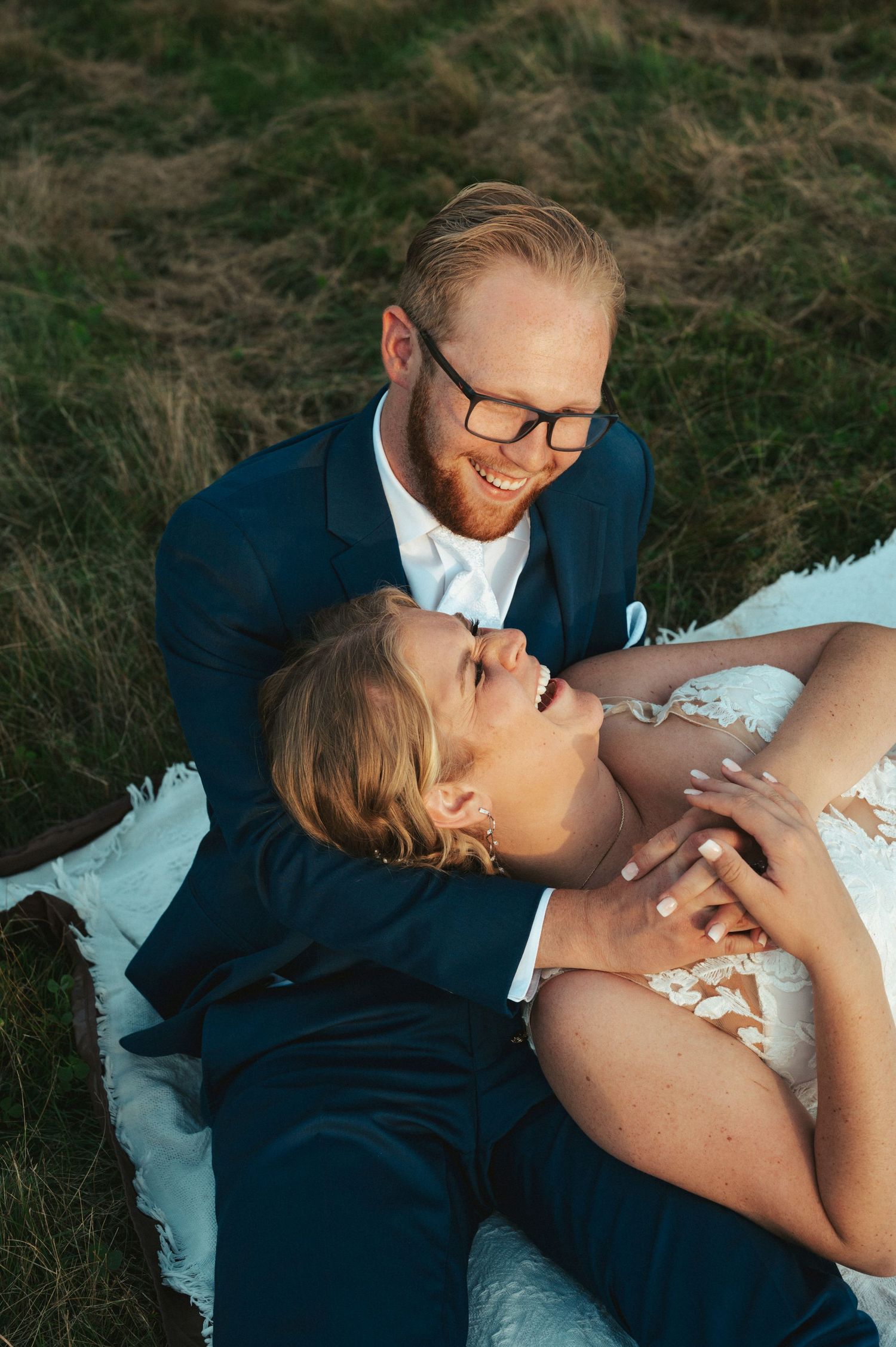 A tender moment between a couple in formal attire laying in tall grass at sunset.