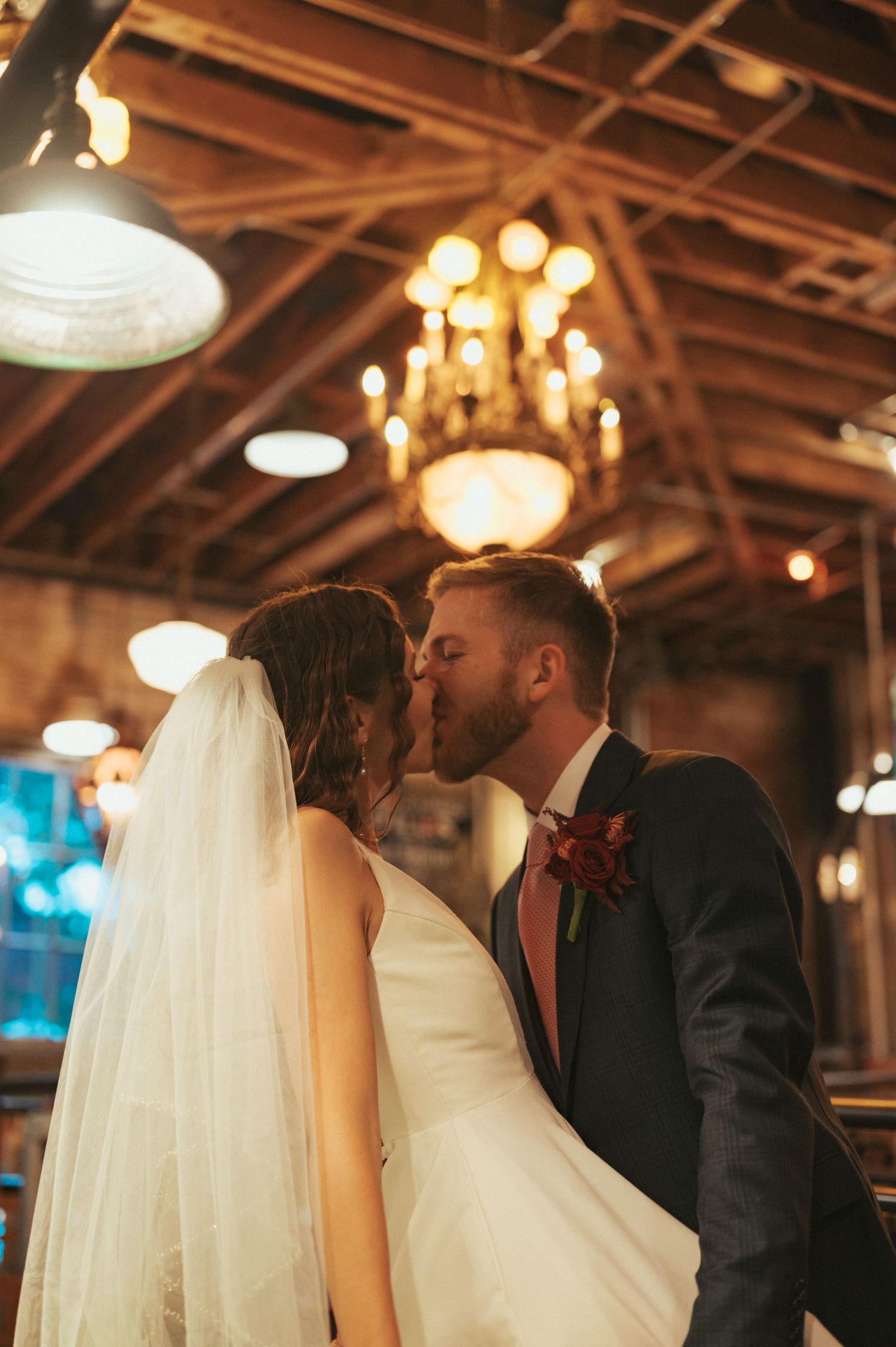 A wedding kiss at an indoor venue with warm lighting and a chandelier in the background.